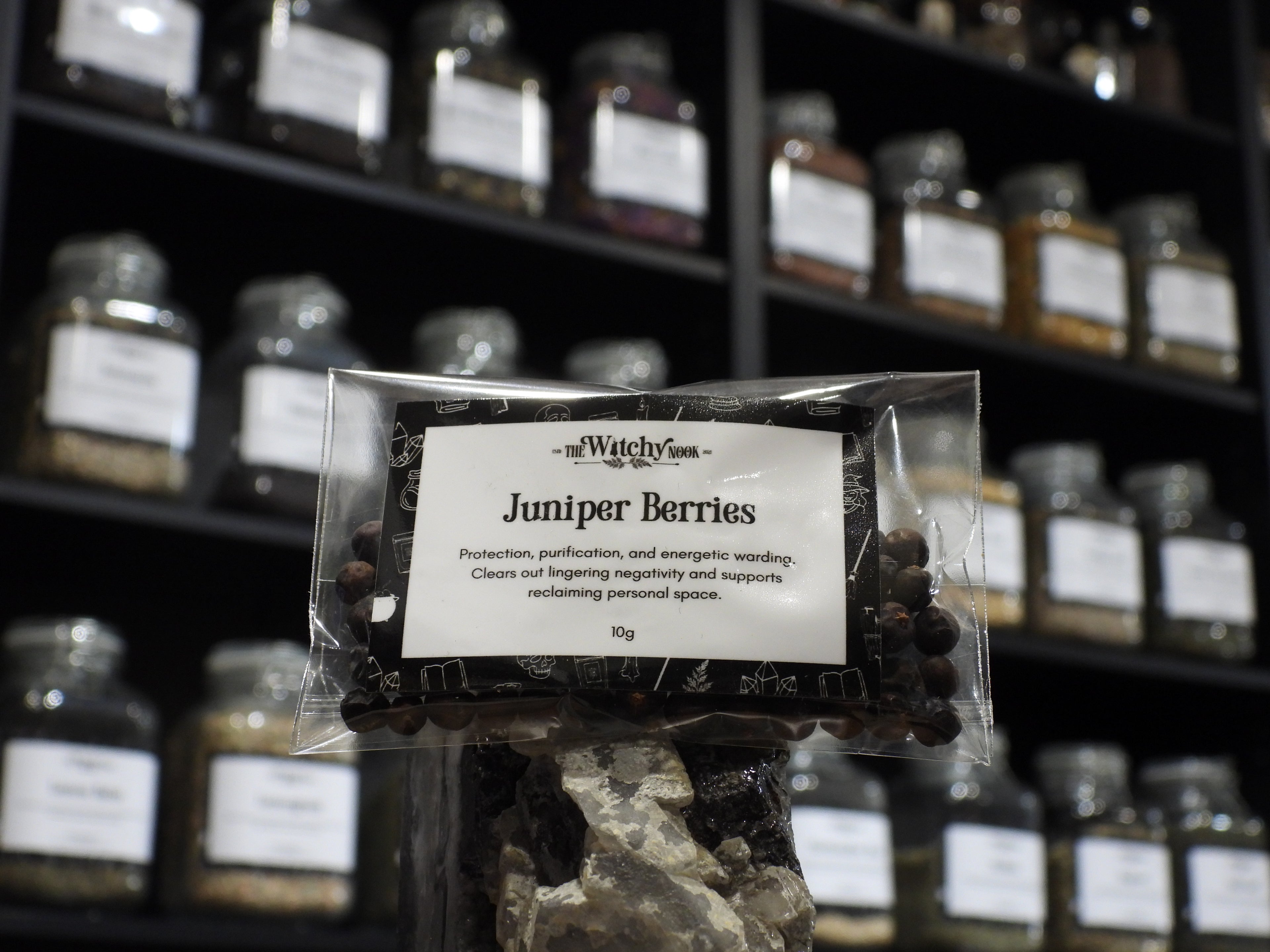 Packaged product labelled ‘Juniper Berries' by 'Witchy Nook' sitting on a crystal with an apothecary of herb jars blurred in the background.