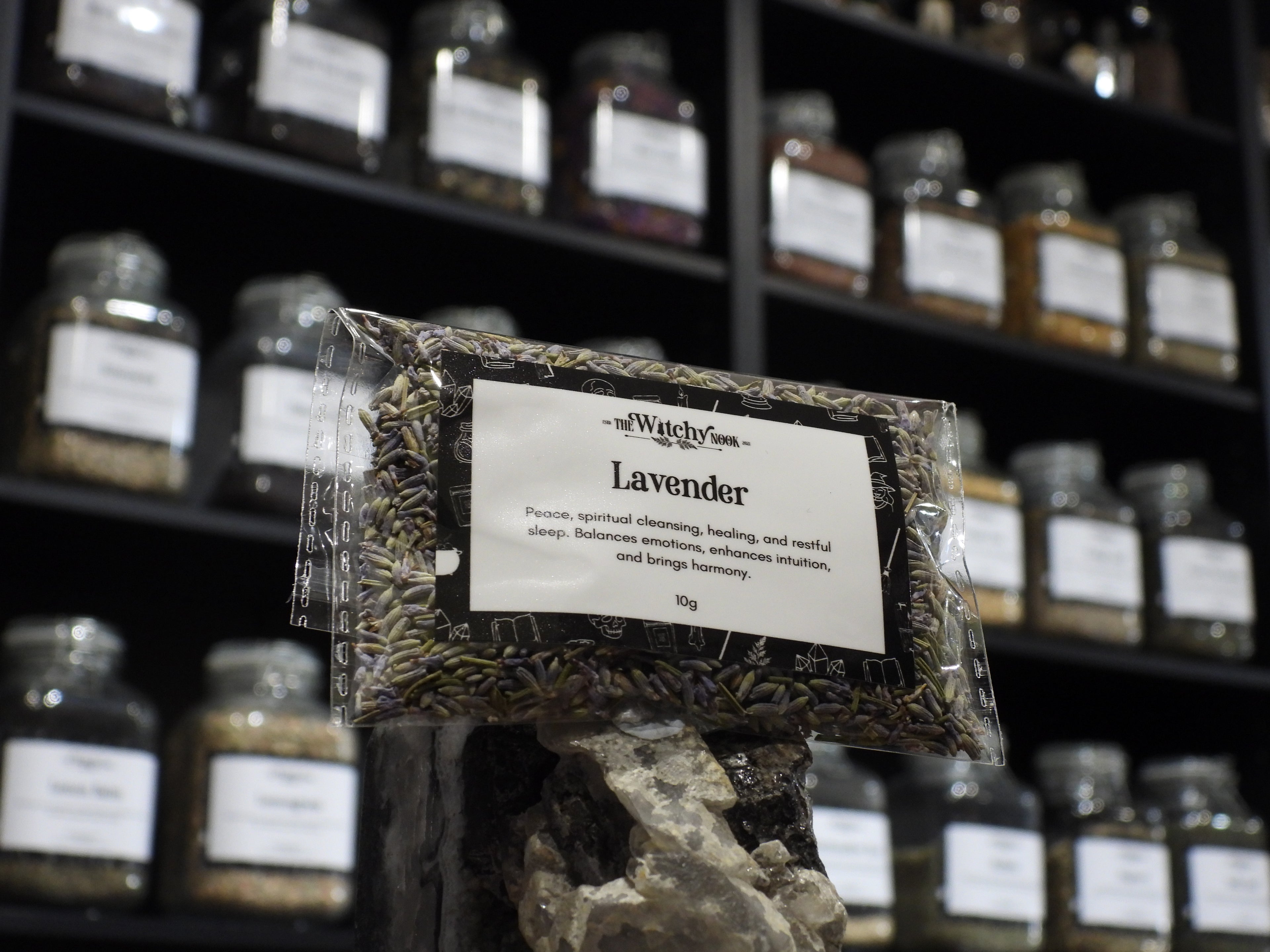 Packaged product labelled ‘Lavender' by 'Witchy Nook' sitting on a crystal with an apothecary of herb jars blurred in the background.