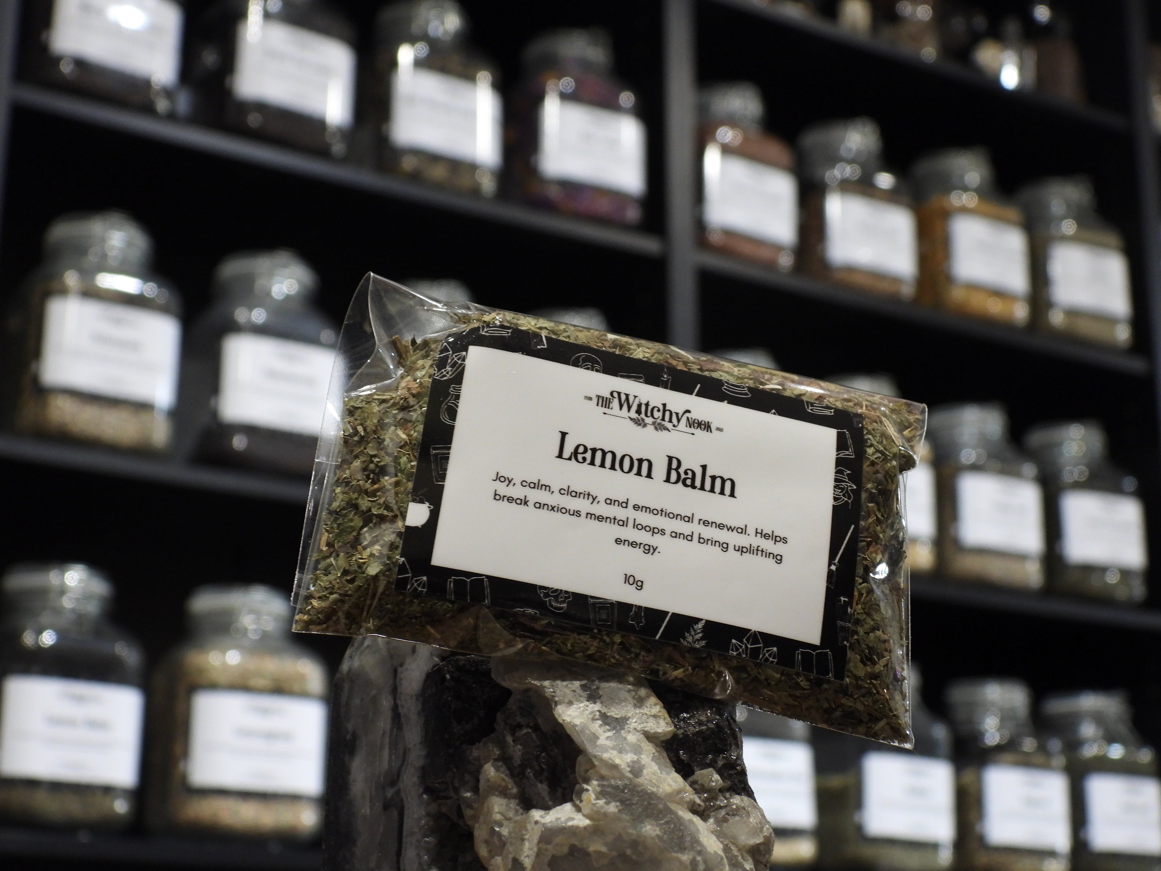 Packaged product labelled ‘Lemon Balm' by 'Witchy Nook' sitting on a crystal with an apothecary of herb jars blurred in the background.