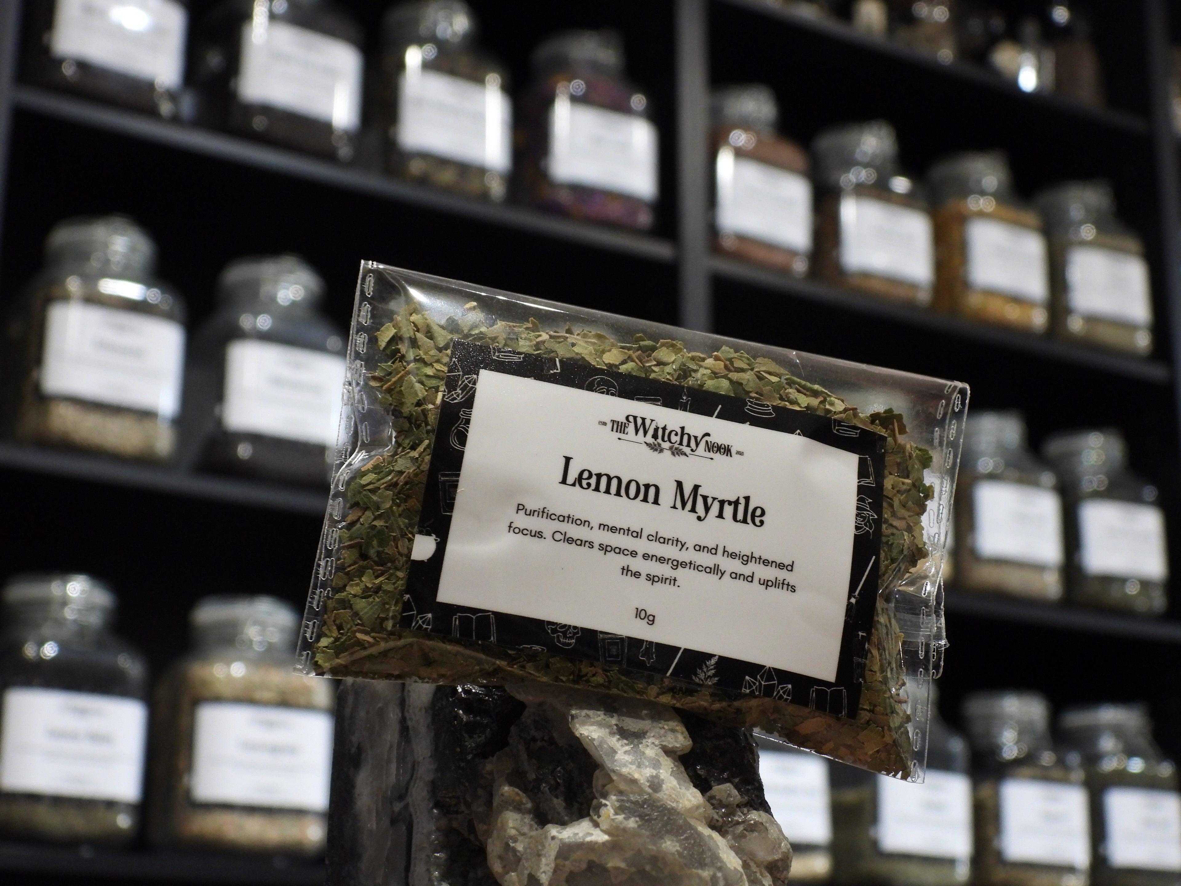 Packaged product labelled ‘Lemon Myrtle' by 'Witchy Nook' sitting on a crystal with an apothecary of herb jars blurred in the background.