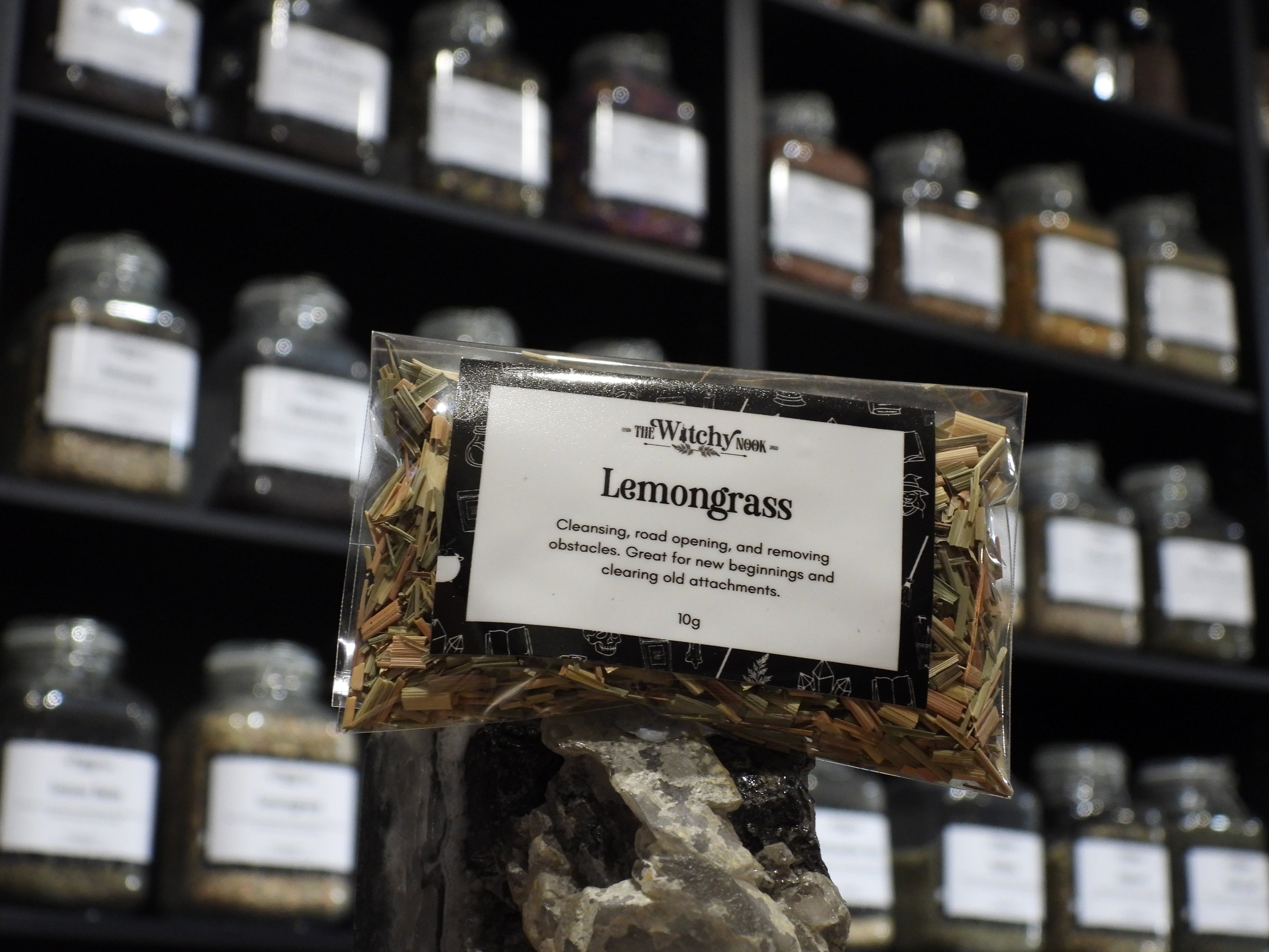 Packaged product labelled ‘Lemongrass' by 'Witchy Nook' sitting on a crystal with an apothecary of herb jars blurred in the background.