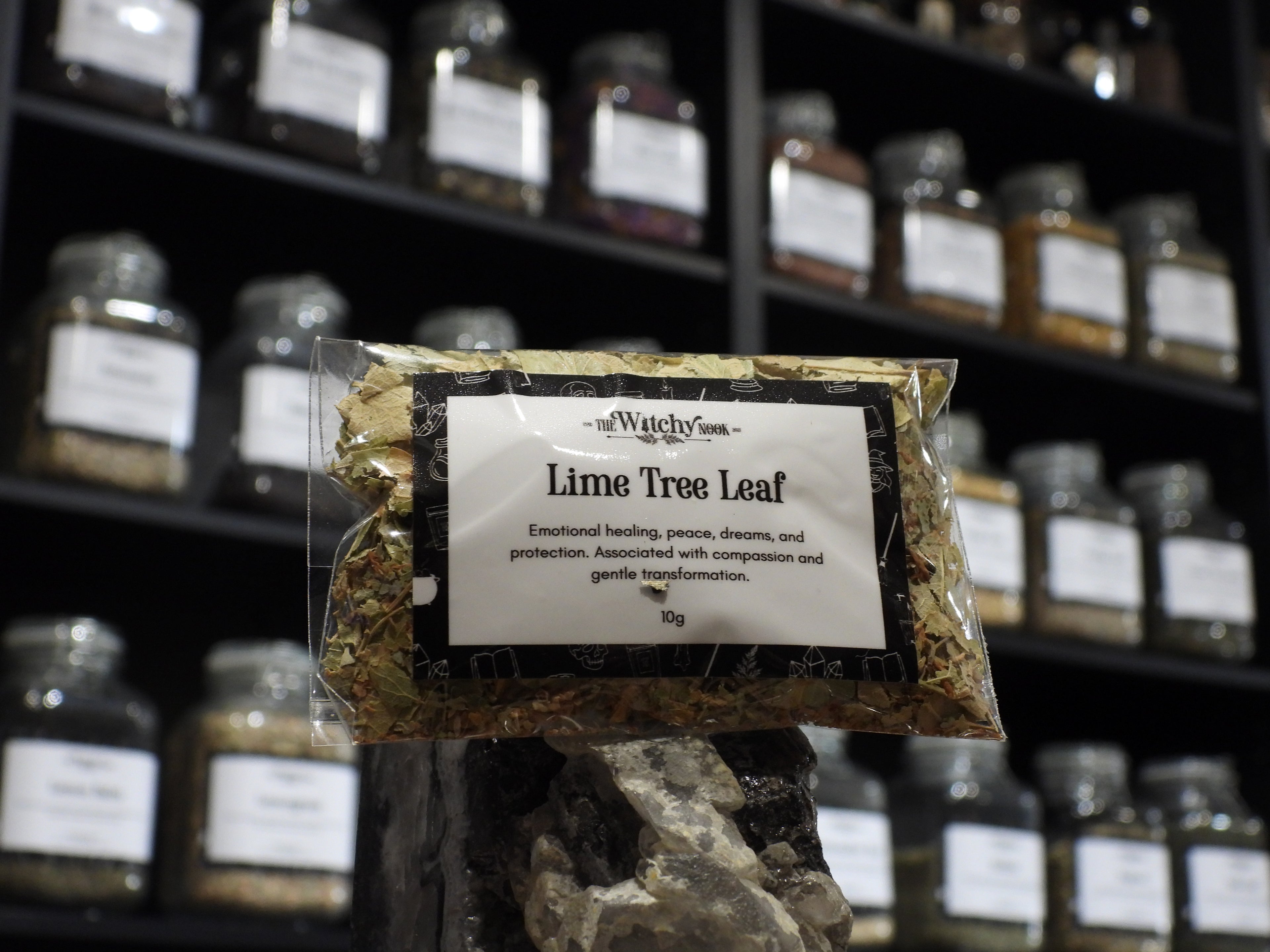 Packaged product labelled ‘Lime Tree Leaf' by 'Witchy Nook' sitting on a crystal with an apothecary of herb jars blurred in the background.