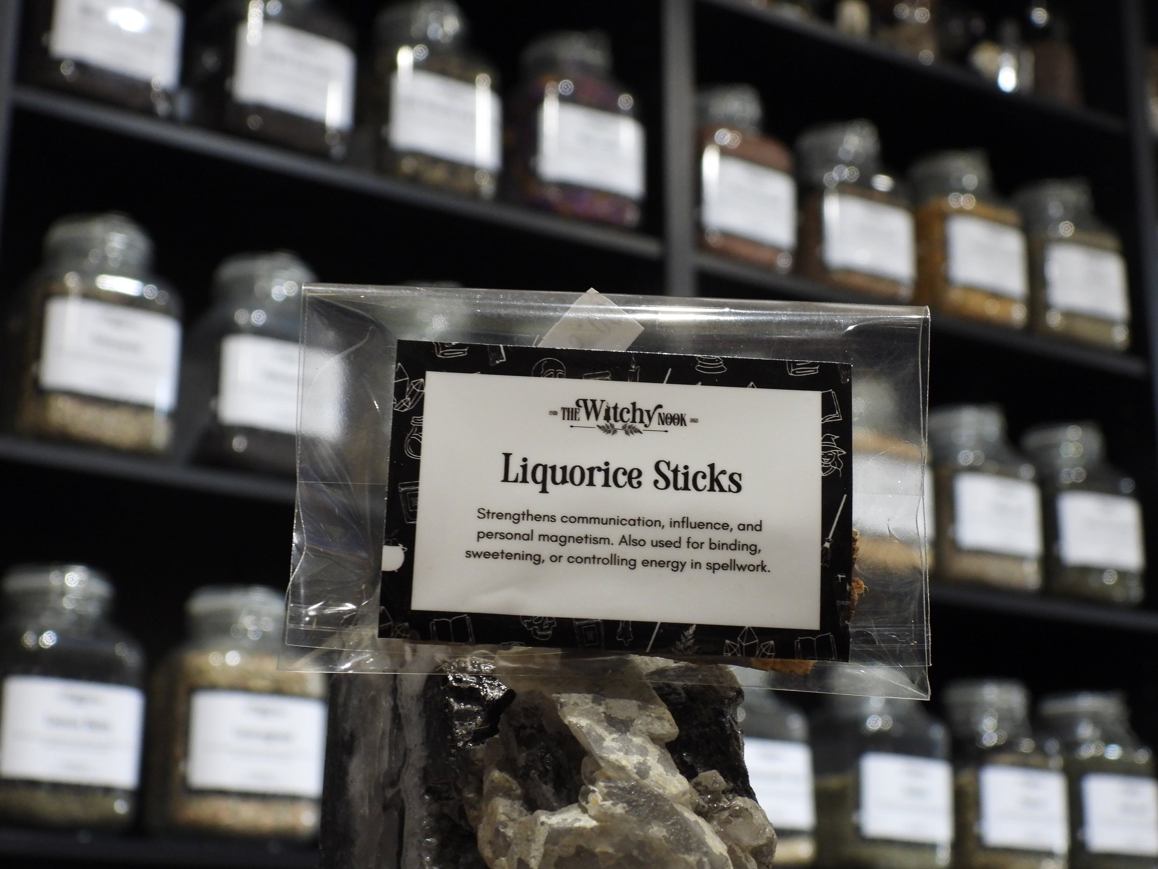 Packaged product labelled ‘Liquorice Sticks' by 'Witchy Nook' sitting on a crystal with an apothecary of herb jars blurred in the background.