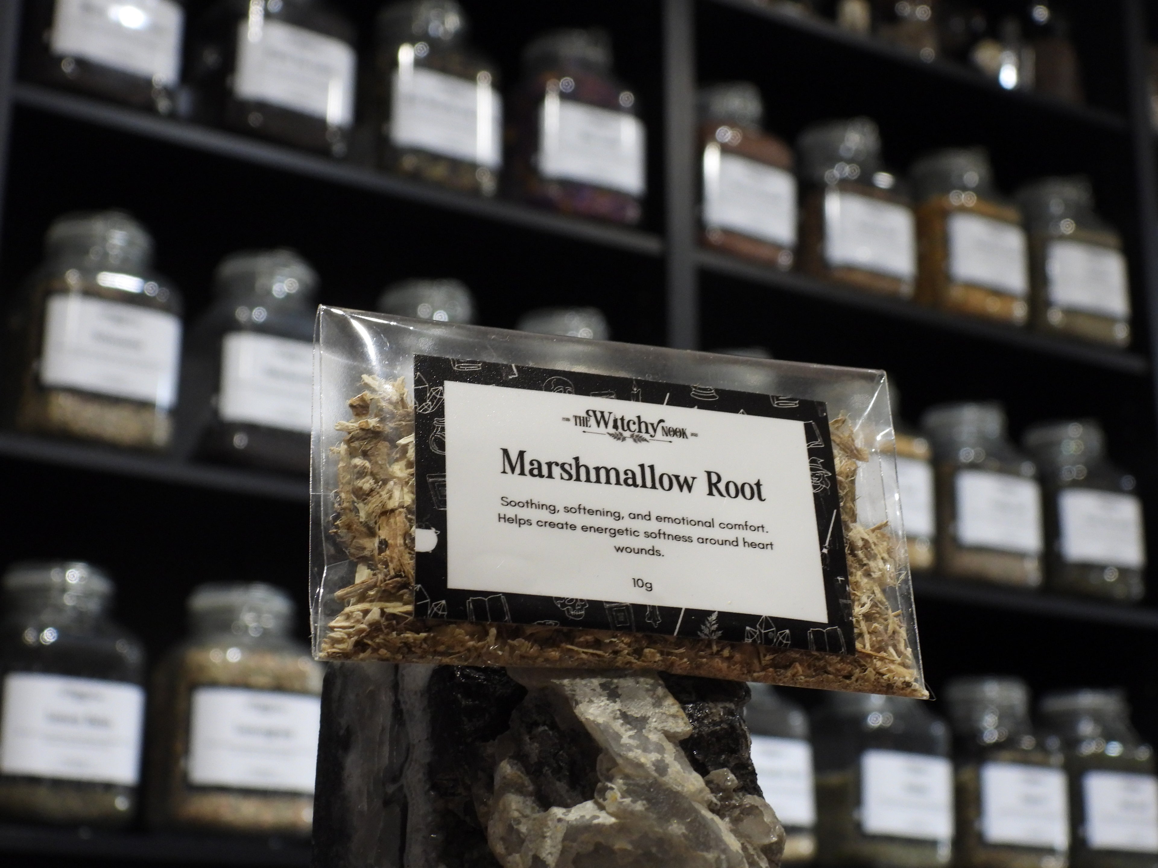 Packaged product labelled ‘Chilli Flakes' by 'Witchy Nook' sitting on a crystal with an apothecary of herb jars blurred in the background.