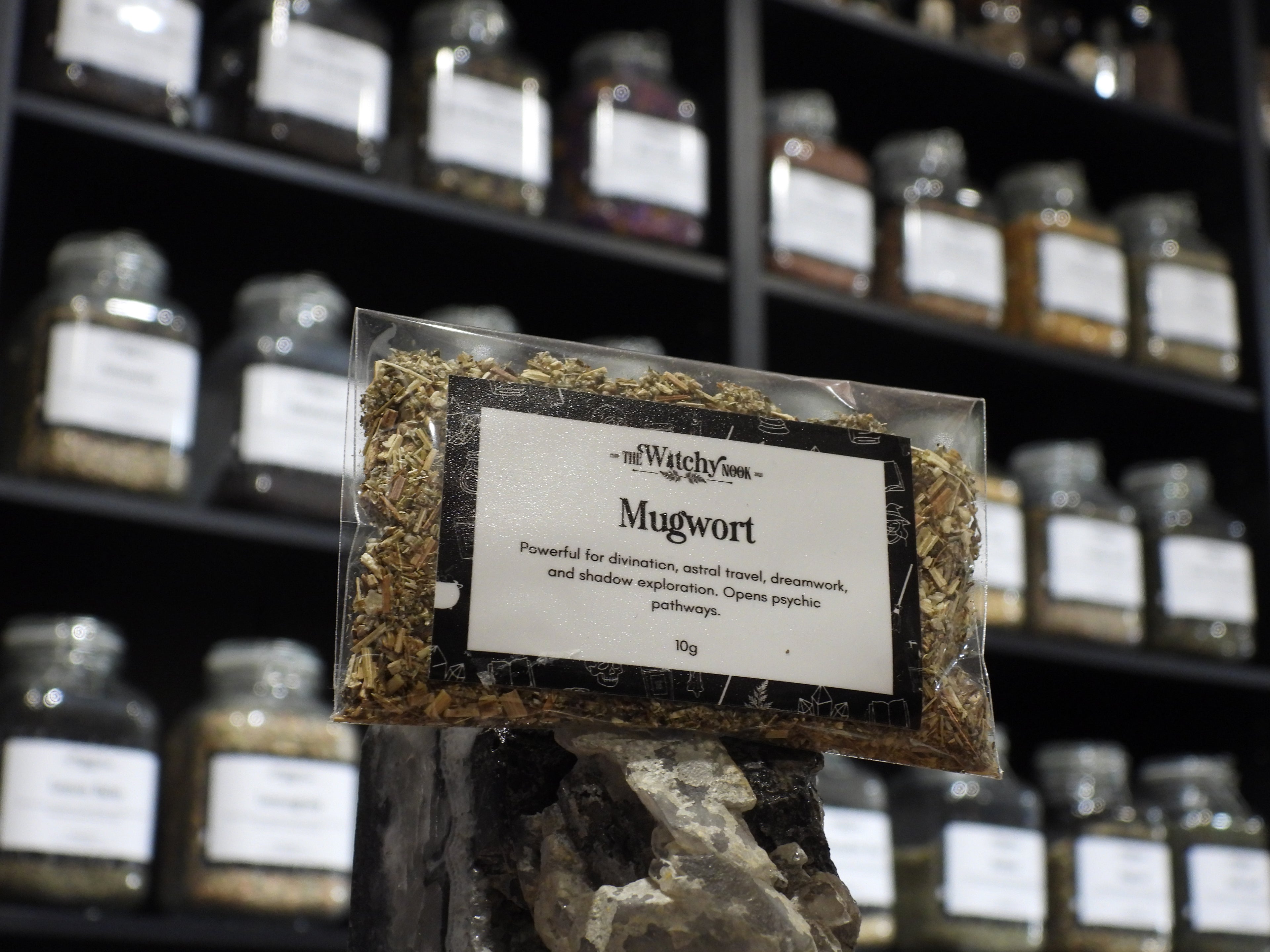 Packaged product labelled ‘Mugwort' by 'Witchy Nook' sitting on a crystal with an apothecary of herb jars blurred in the background.