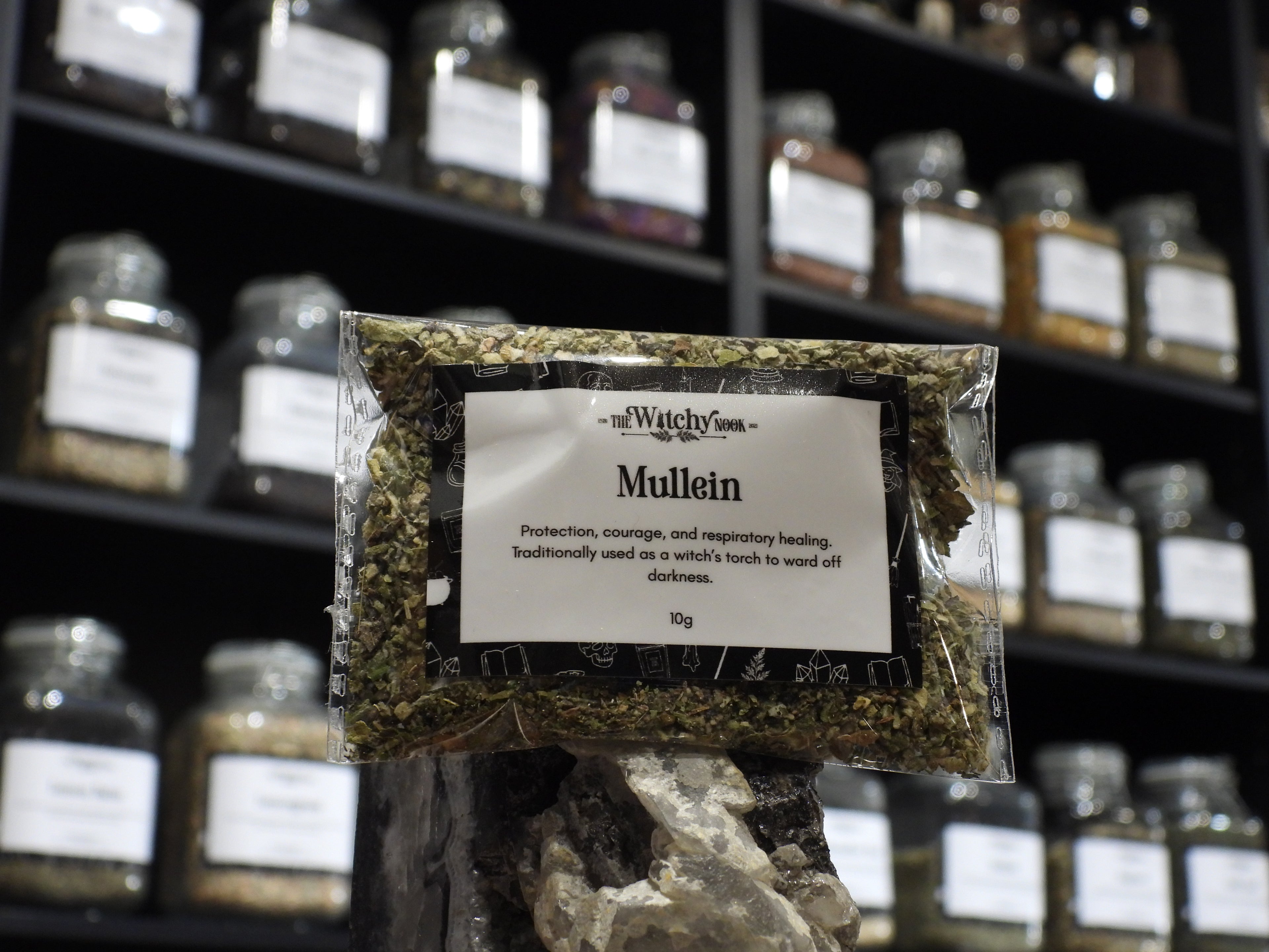 Packaged product labelled ‘Mullein’ by 'The Witchy Nook' sitting on a crystal with an apothecary of herb jars blurred in the background.