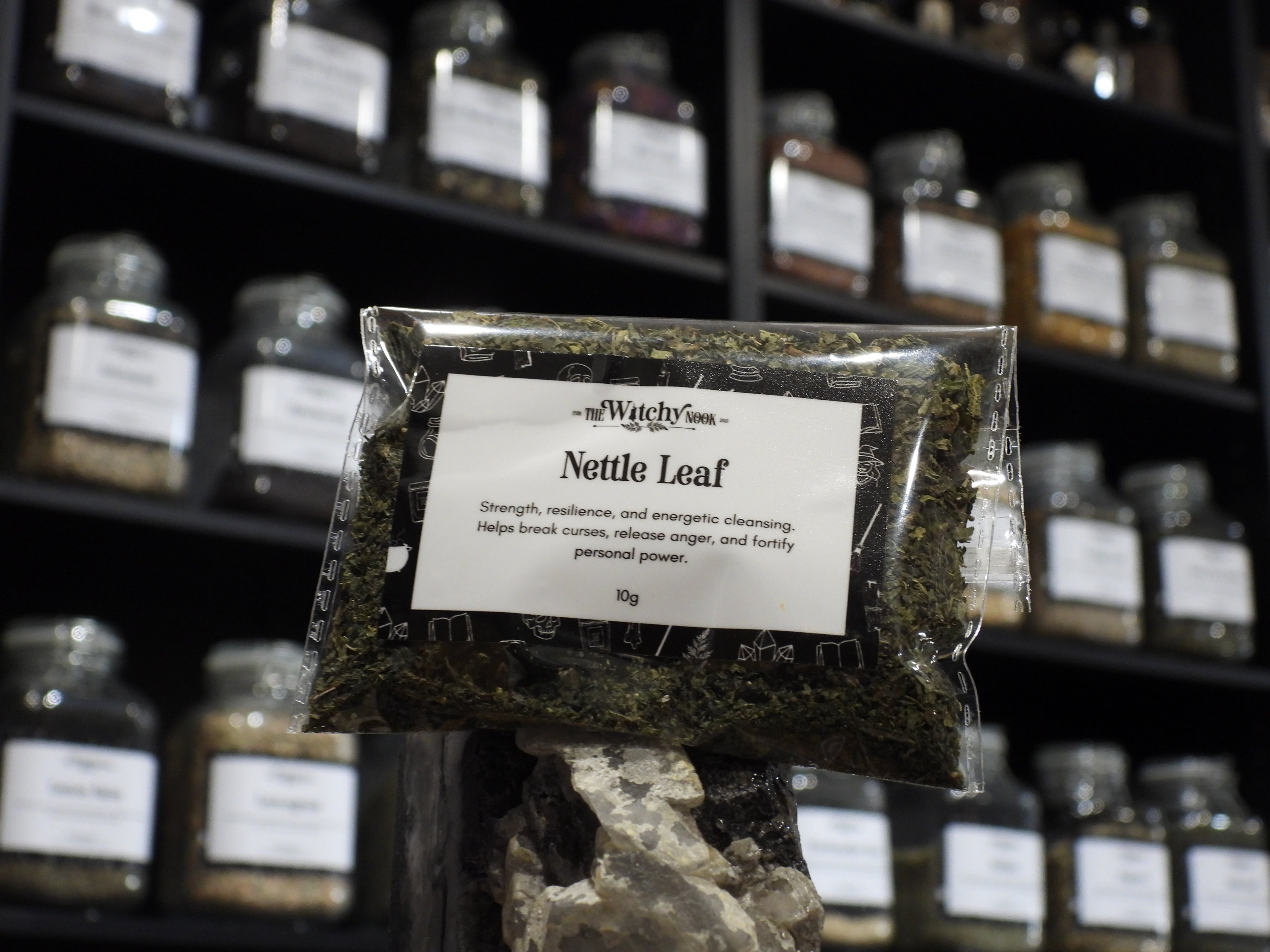 Packaged product labelled ‘Nettle Leaf' by 'Witchy Nook' sitting on a crystal with an apothecary of herb jars blurred in the background.