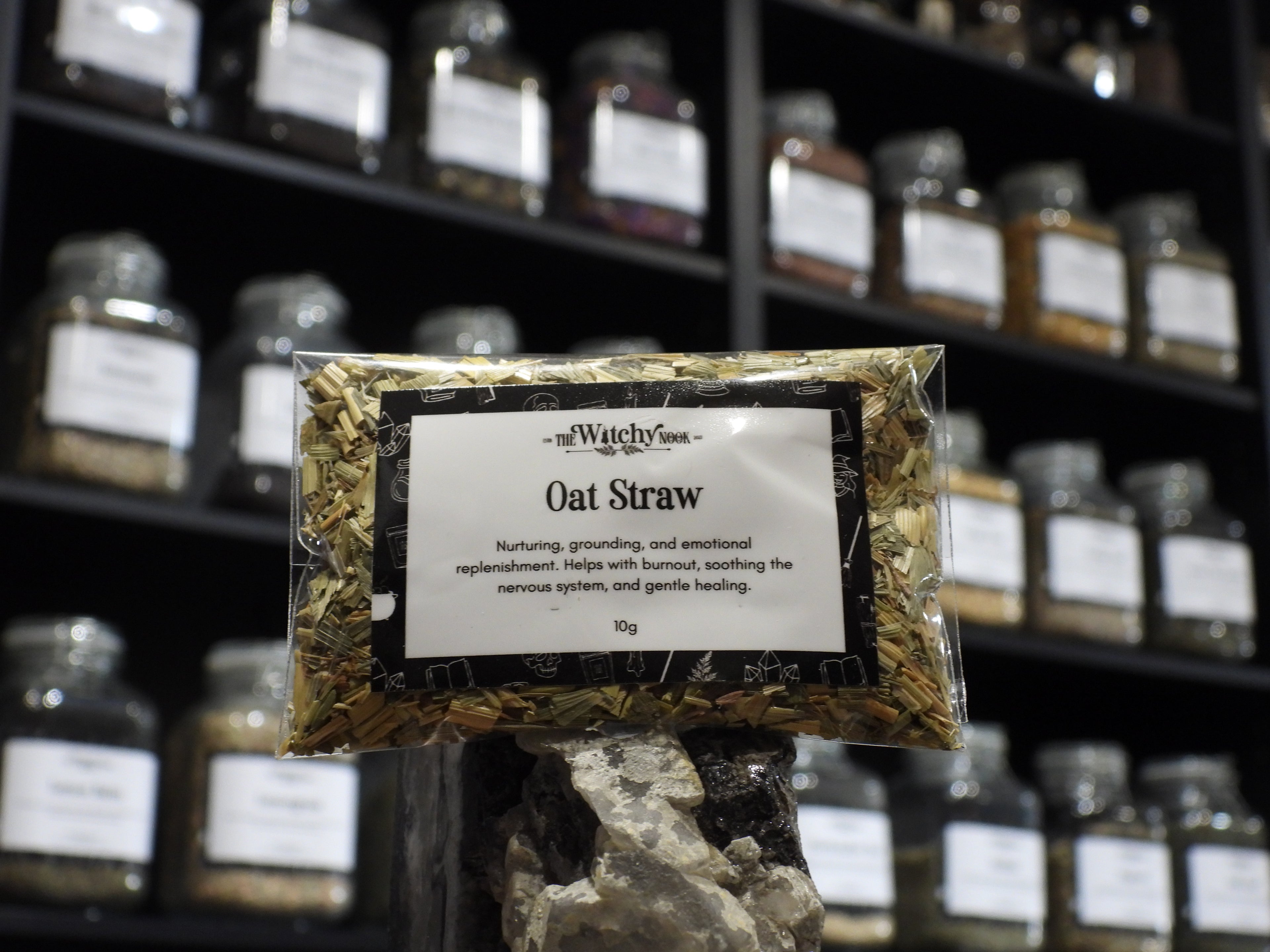 Packaged product labelled ‘Oat Straw’ by 'The Witchy Nook' sitting on a crystal with an apothecary of herb jars blurred in the background.