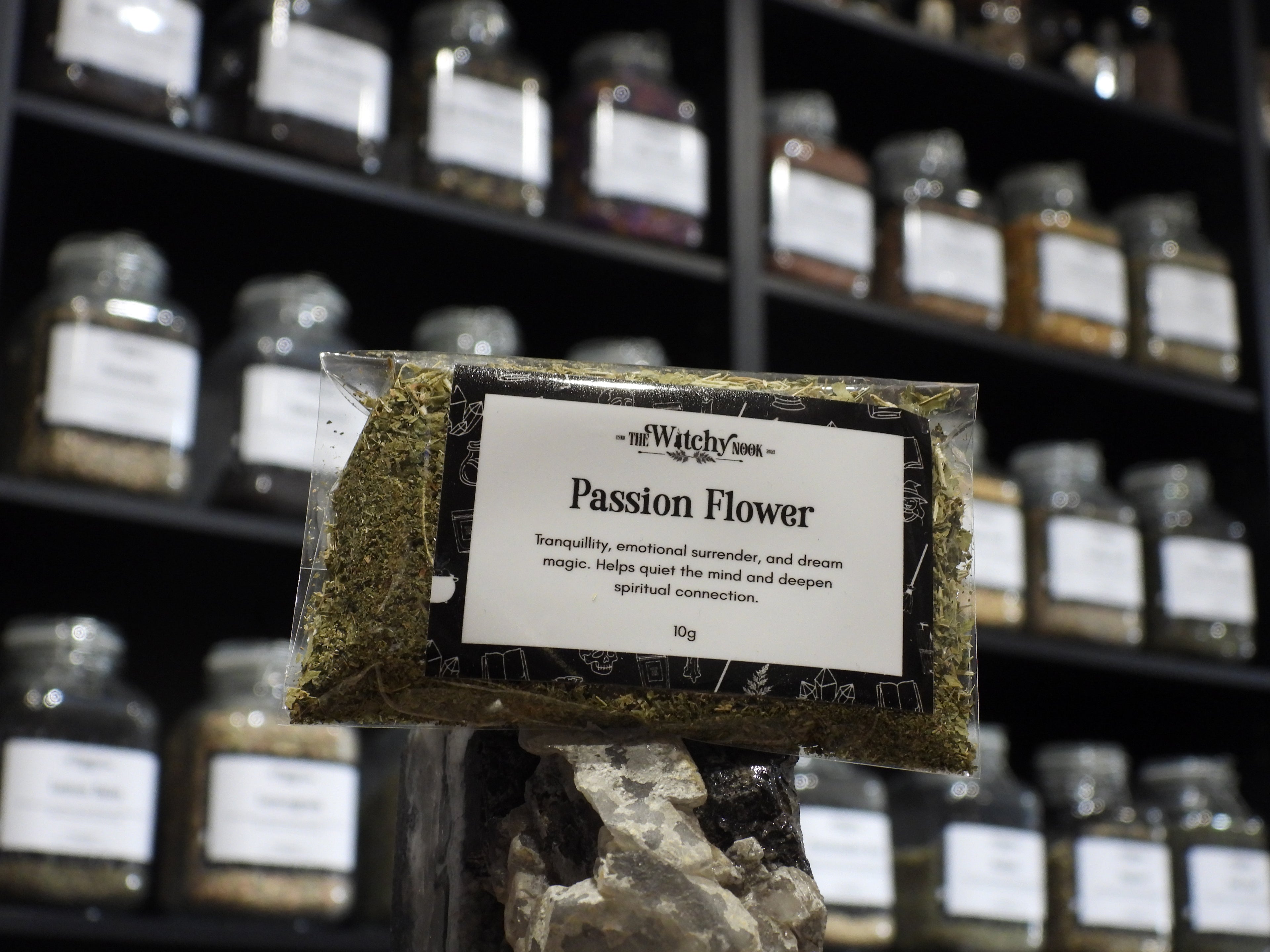 Packaged product labelled ‘Passion Flower' by 'Witchy Nook' sitting on a crystal with an apothecary of herb jars blurred in the background.