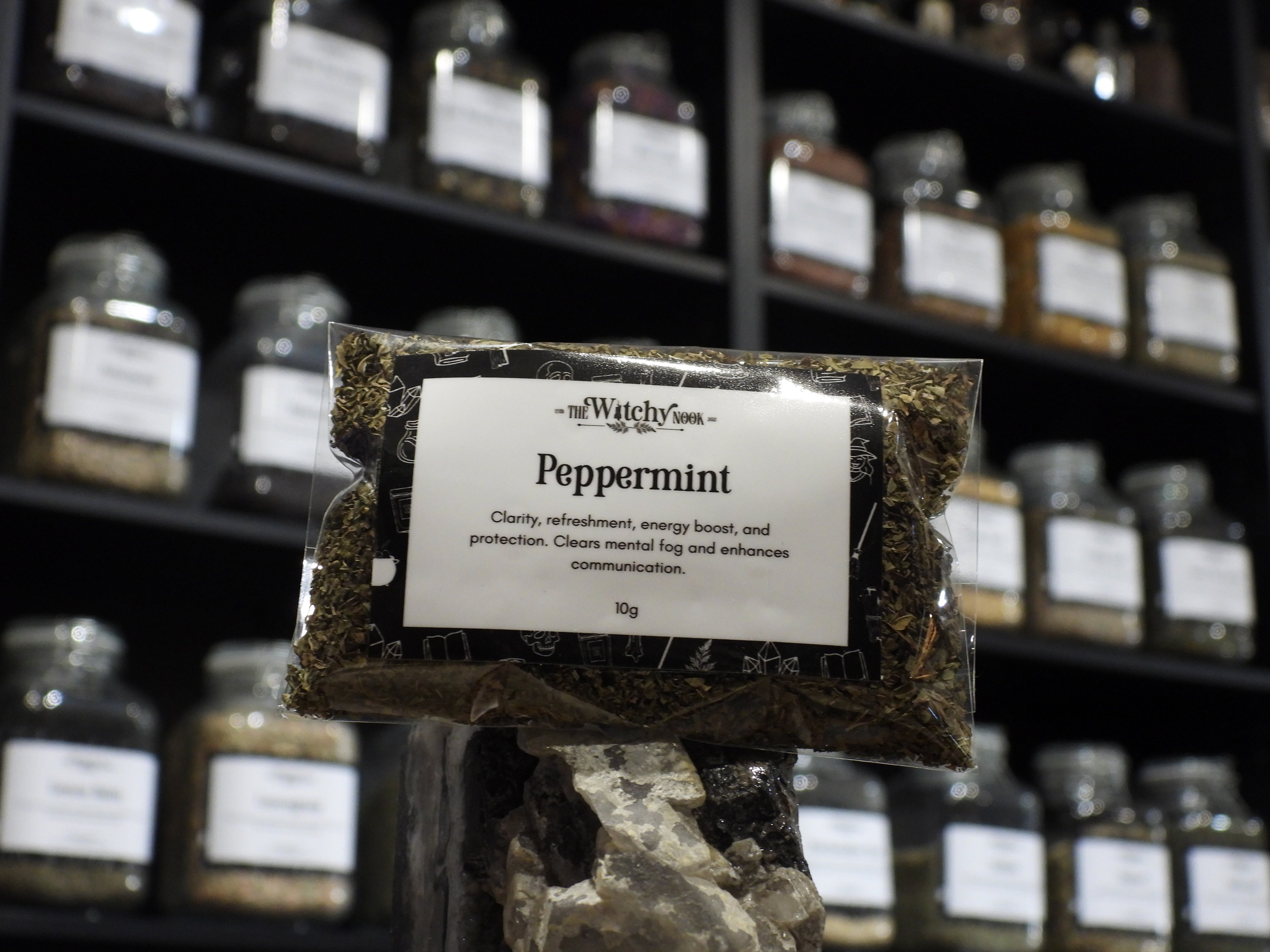 Packaged product labelled ‘Peppermint’ by 'The Witchy Nook' sitting on a crystal with an apothecary of herb jars blurred in the background.