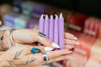 Purple candles held by a person with tattoos and rings in a blurred store setting