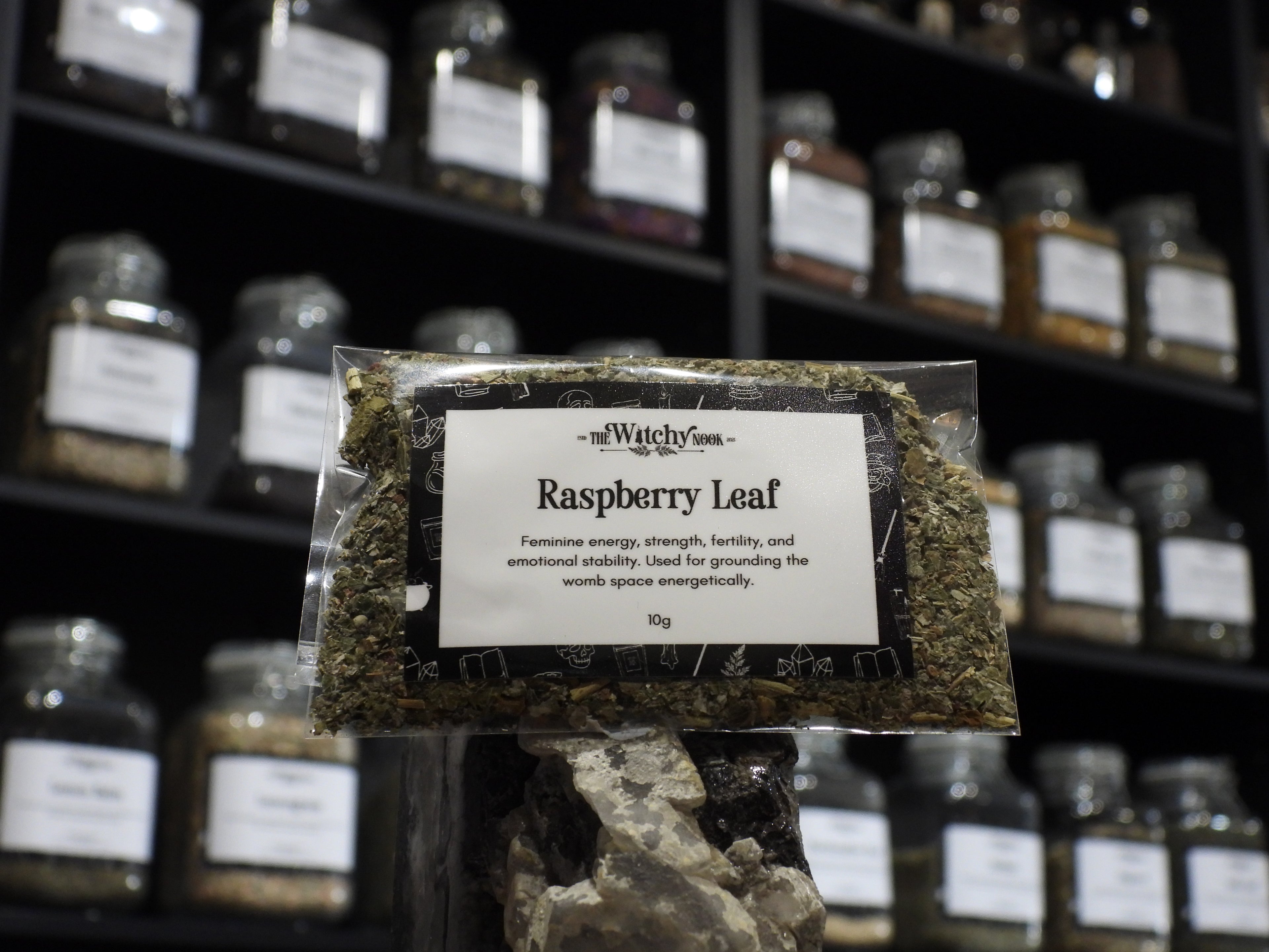 Packaged product labelled ‘Raspberry Leaf' by 'Witchy Nook' sitting on a crystal with an apothecary of herb jars blurred in the background.