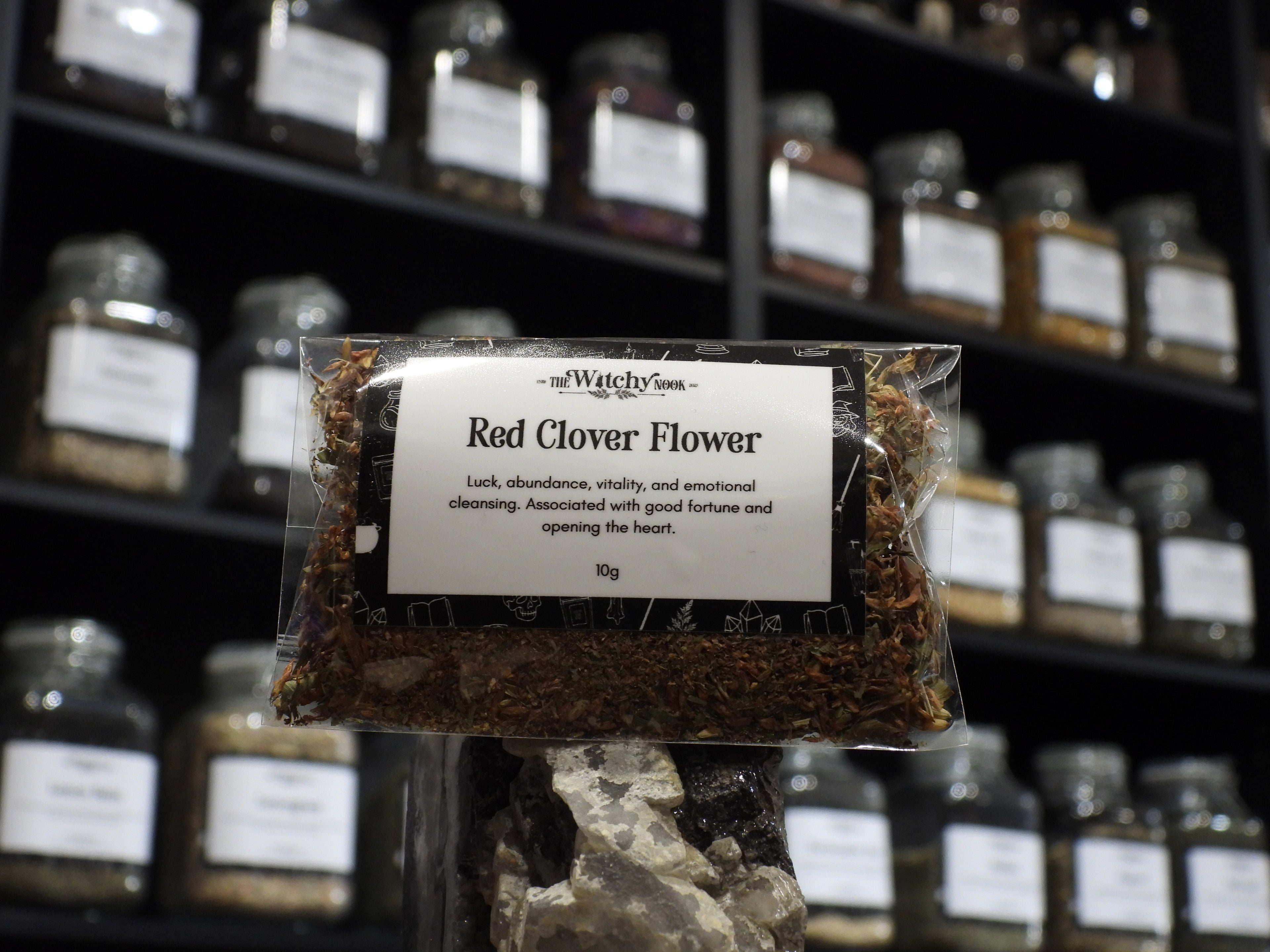 Packaged product labelled ‘Red Clover Flower' by 'Witchy Nook' sitting on a crystal with an apothecary of herb jars blurred in the background.