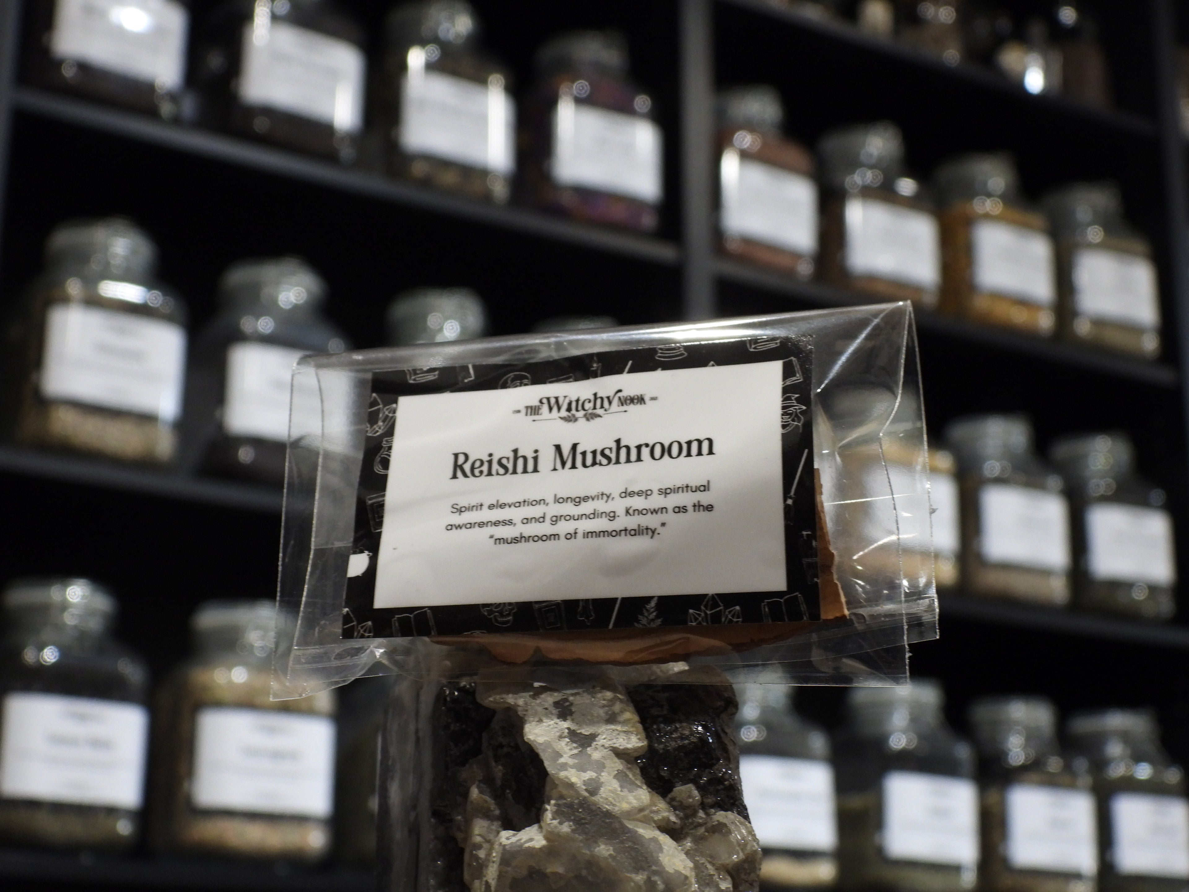 Packaged product labelled ‘Reishi Mushroom' by 'Witchy Nook' sitting on a crystal with an apothecary of herb jars blurred in the background.