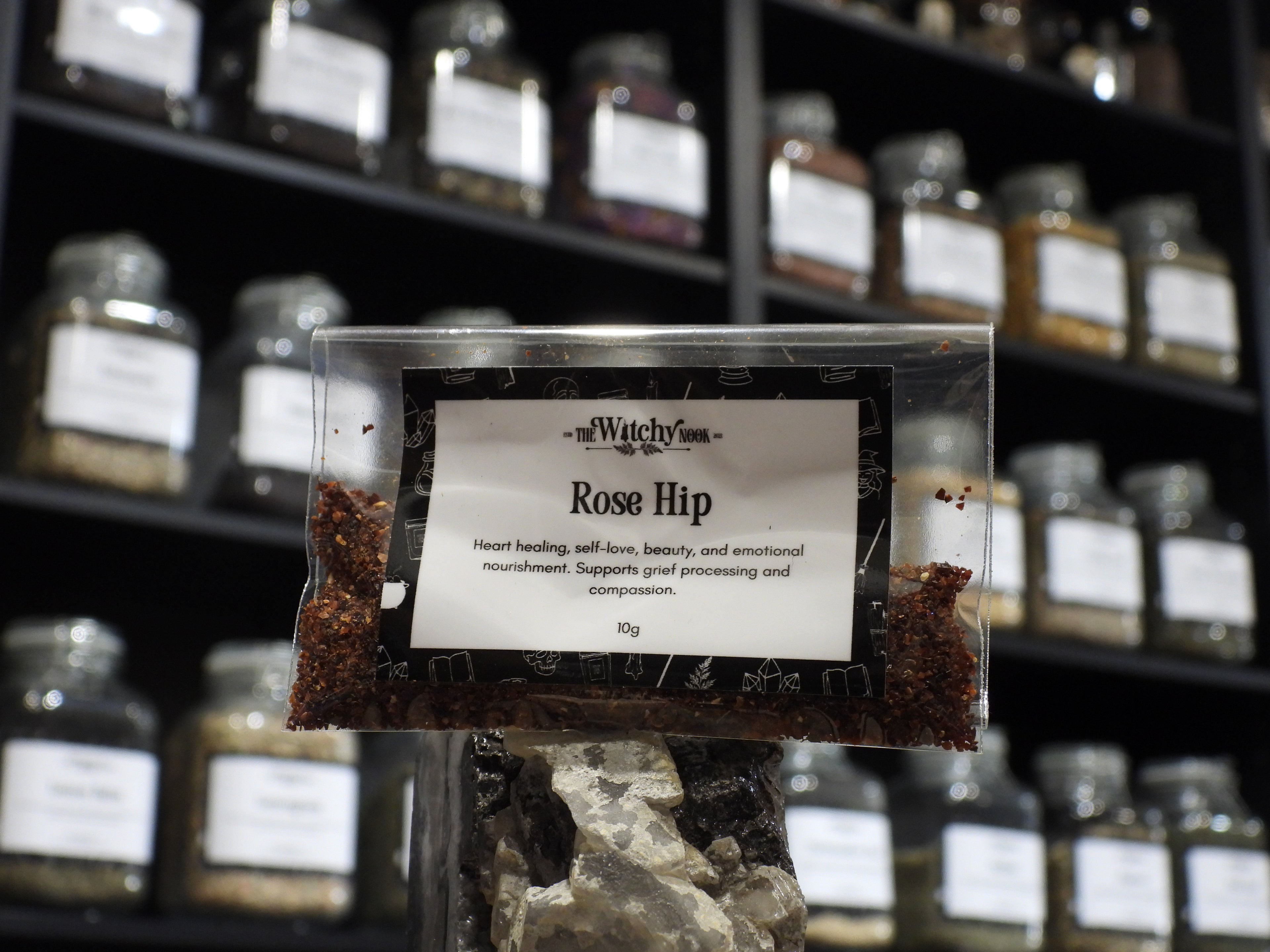 Packaged product labelled ‘Rose Hip' by 'Witchy Nook' sitting on a crystal with an apothecary of herb jars blurred in the background.