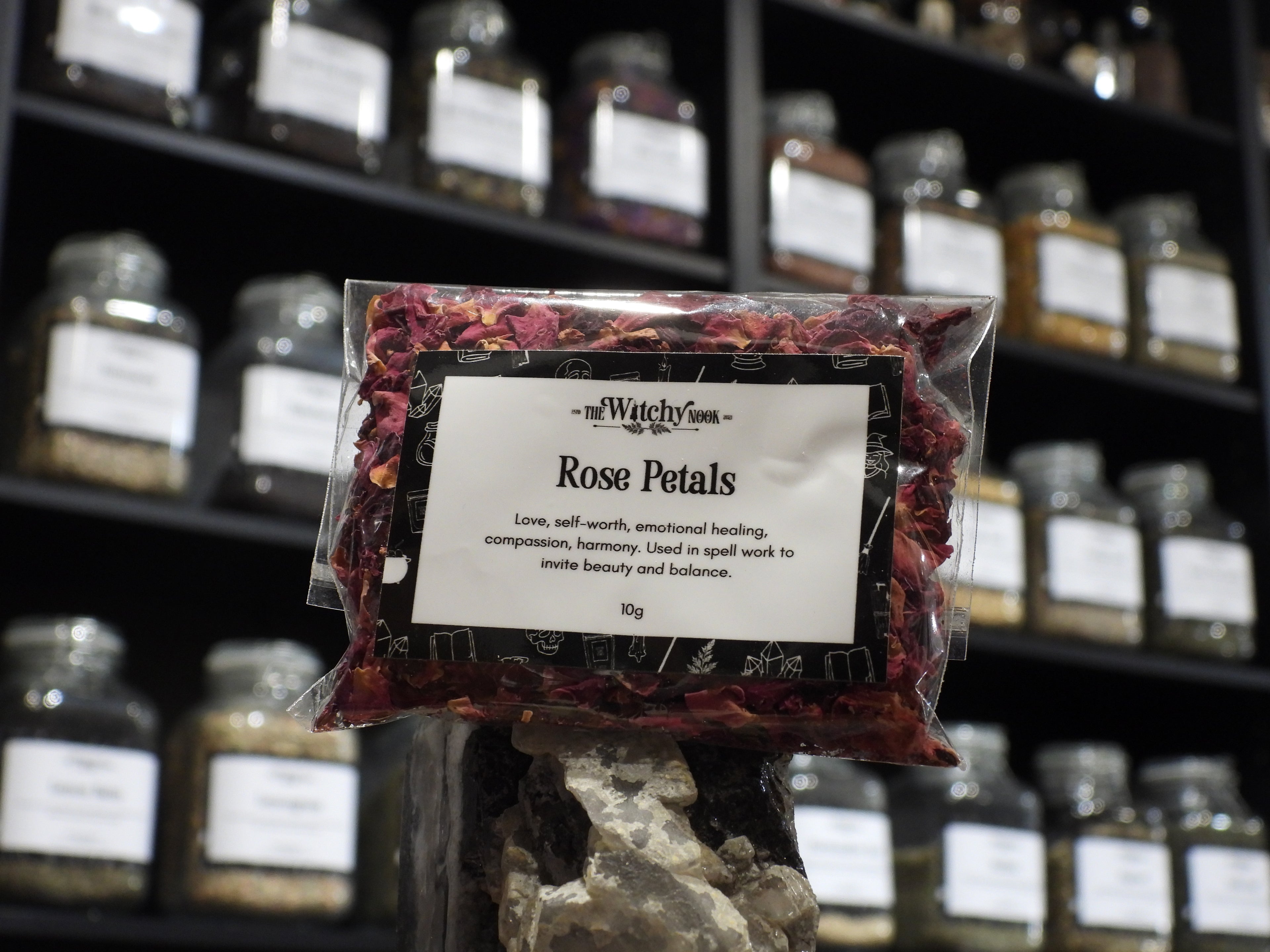 Packaged product labelled ‘Rose Petals' by 'Witchy Nook' sitting on a crystal with an apothecary of herb jars blurred in the background.