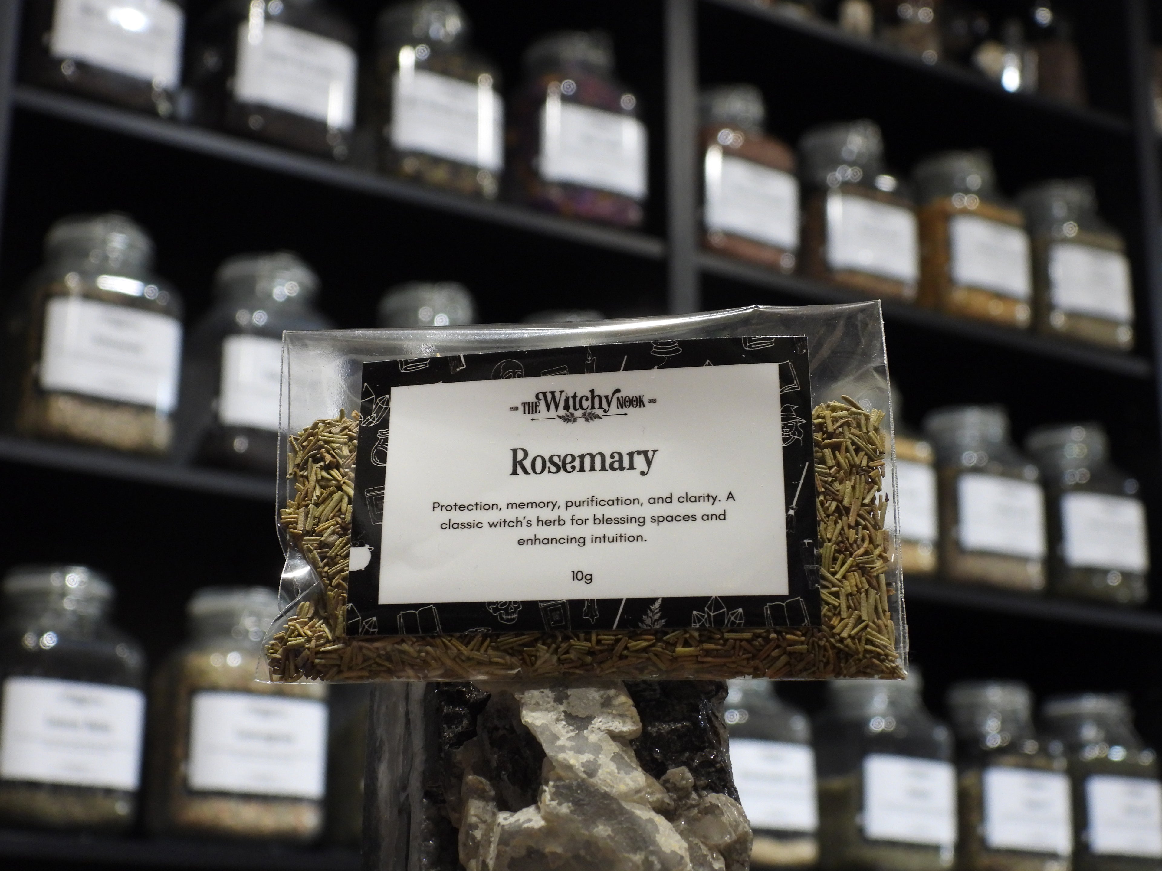 Packaged product labelled ‘Rosemary' by 'Witchy Nook' sitting on a crystal with an apothecary of herb jars blurred in the background.
