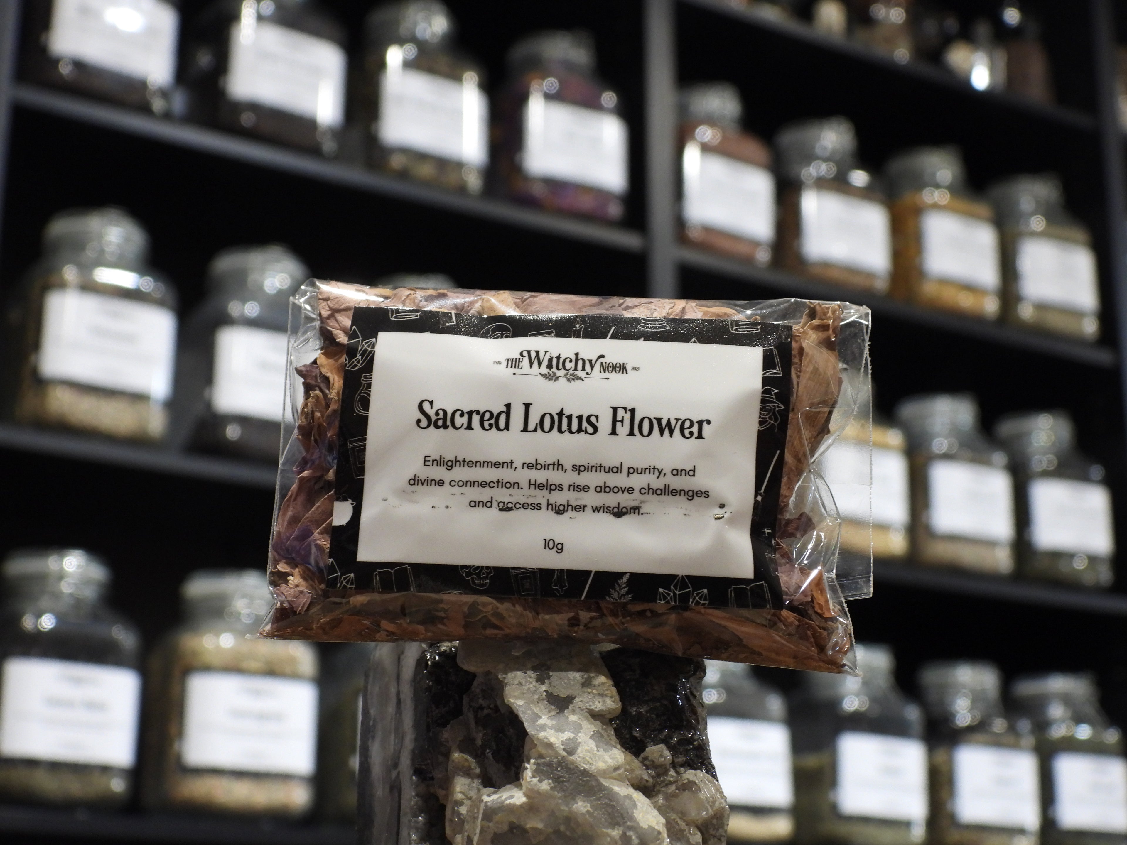 Packaged product labelled ‘Sacred Lotus Flower' by 'Witchy Nook' sitting on a crystal with an apothecary of herb jars blurred in the background.