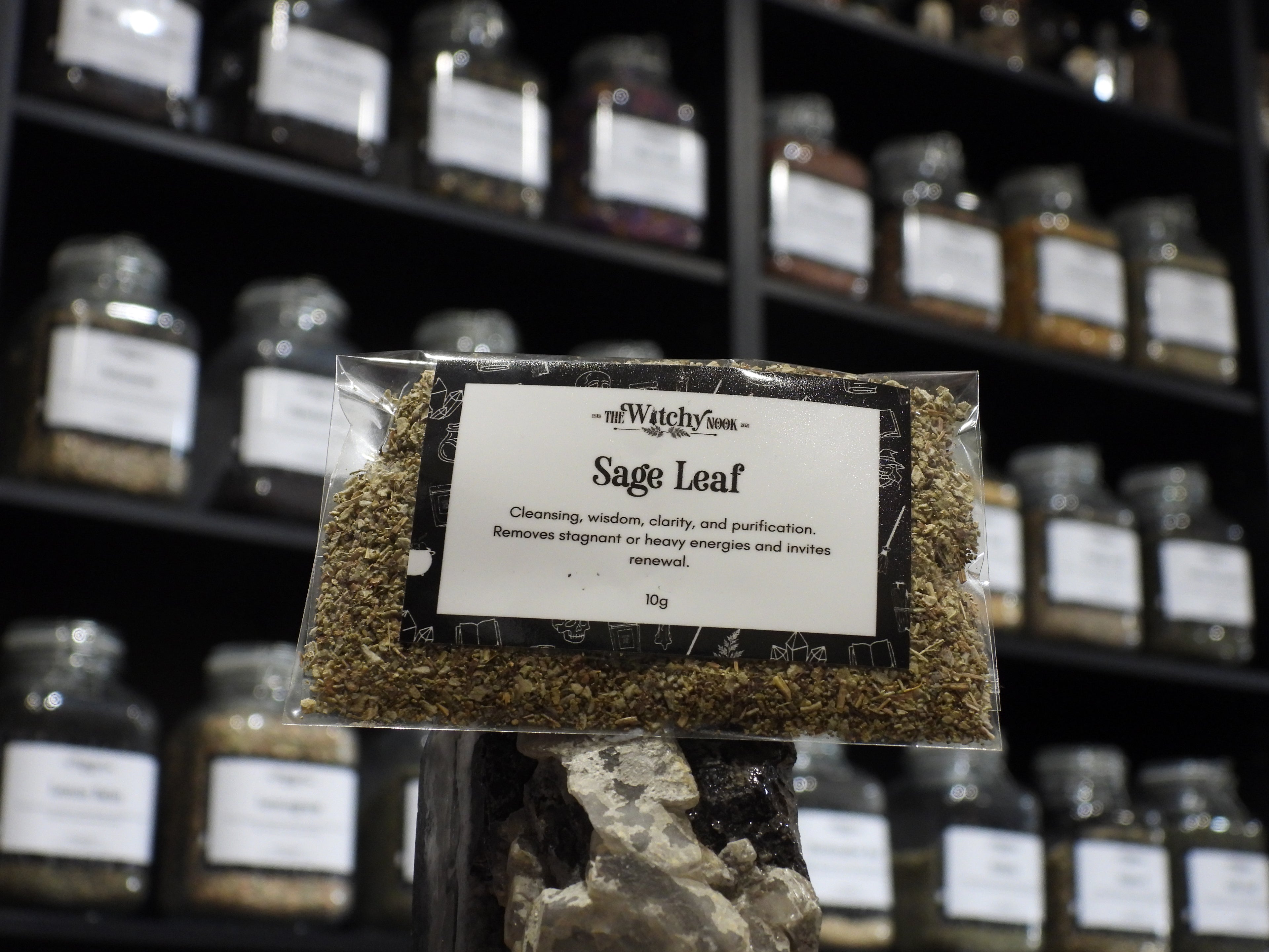 Packaged product labelled ‘Sage leaf’ by 'The Witchy Nook' sitting on a crystal with an apothecary of herb jars blurred in the background.