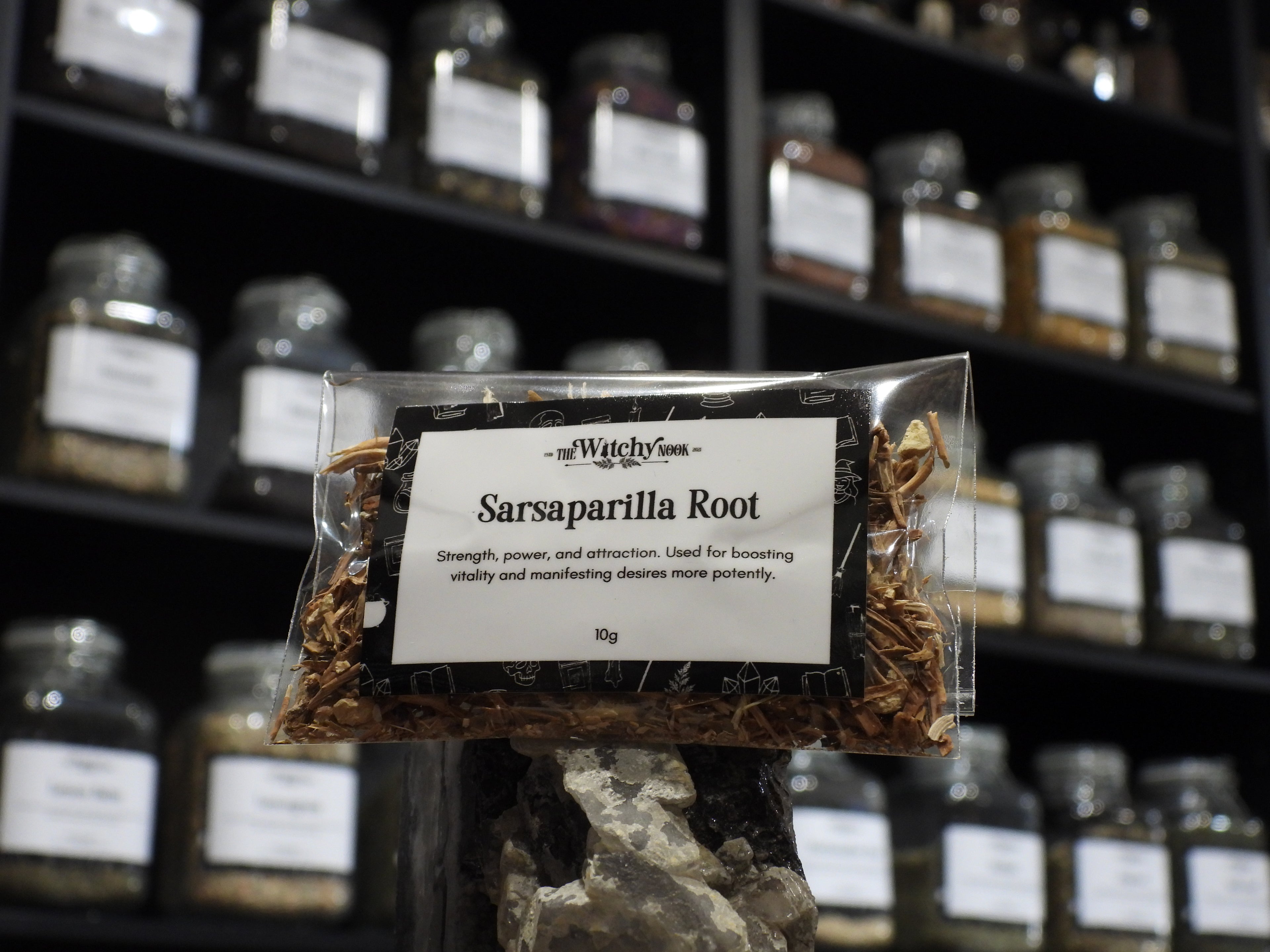 Packaged product labelled ‘Sarsaparilla Root’ by 'The Witchy Nook' sitting on a crystal with an apothecary of herb jars blurred in the background.