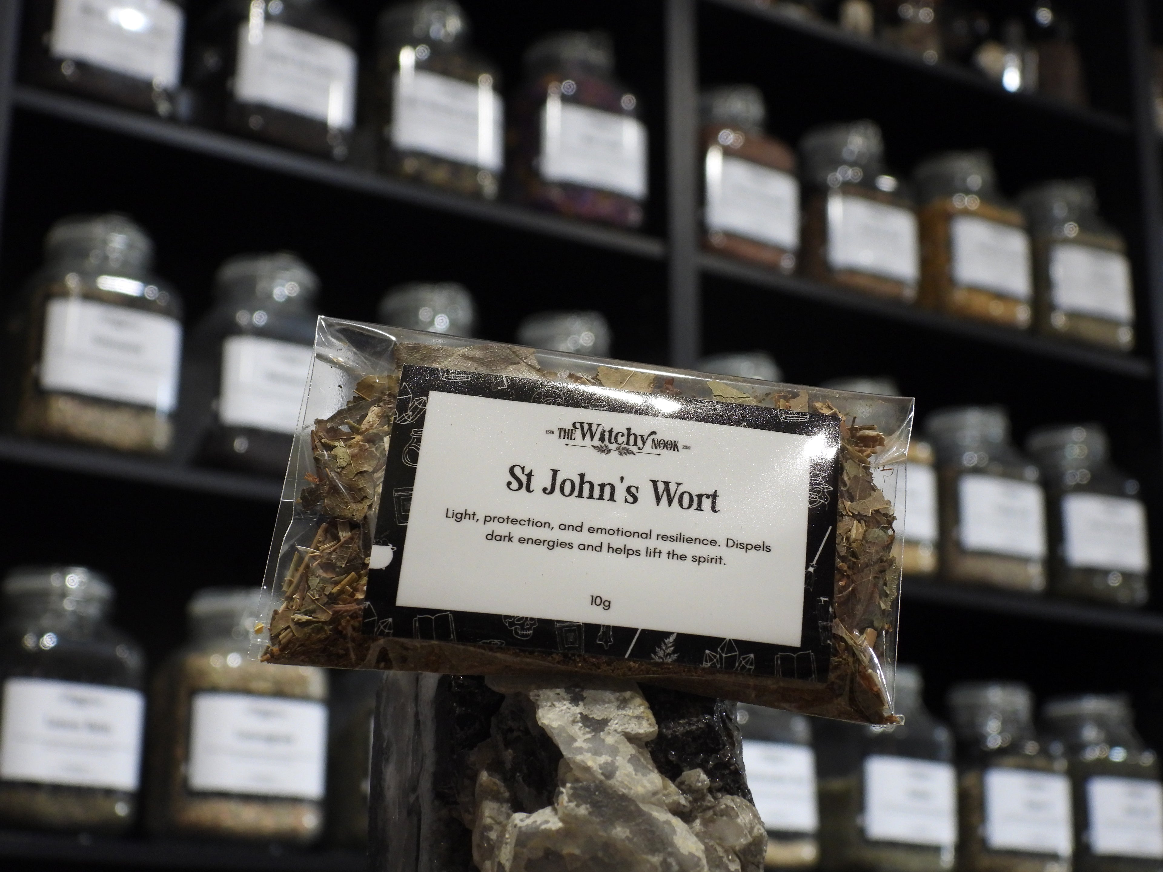 Packaged product labelled ‘St John's Wort' by 'Witchy Nook' sitting on a crystal with an apothecary of herb jars blurred in the background.