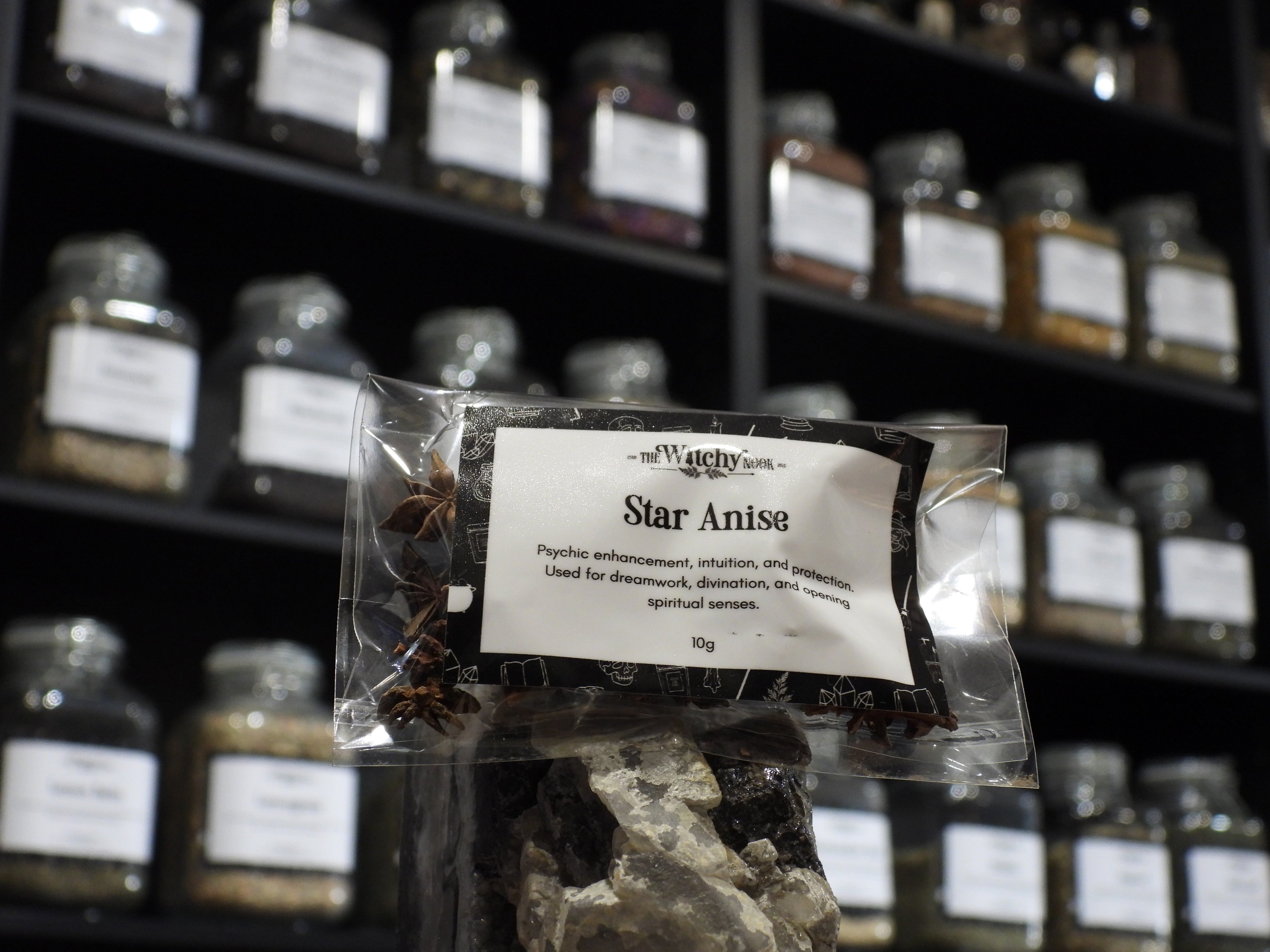 Packaged product labelled ‘Star Anise' by 'Witchy Nook' sitting on a crystal with an apothecary of herb jars blurred in the background.