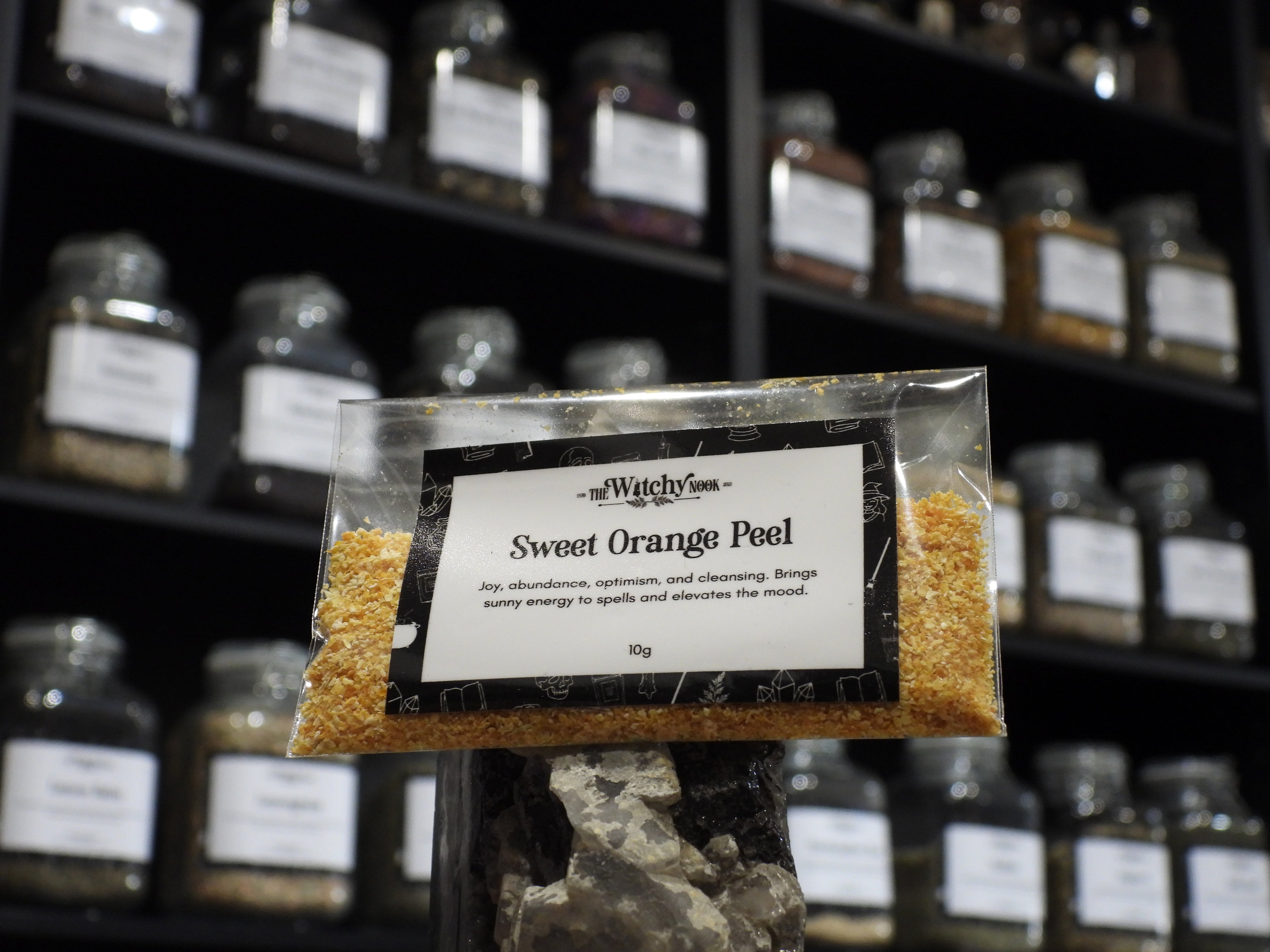 Packaged product labelled ‘Sweet Orange Peel' by 'Witchy Nook' sitting on a crystal with an apothecary of herb jars blurred in the background.
