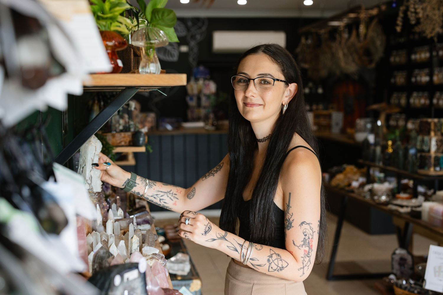 Woman with tattoos and glasses standing in a store with shelves and products.