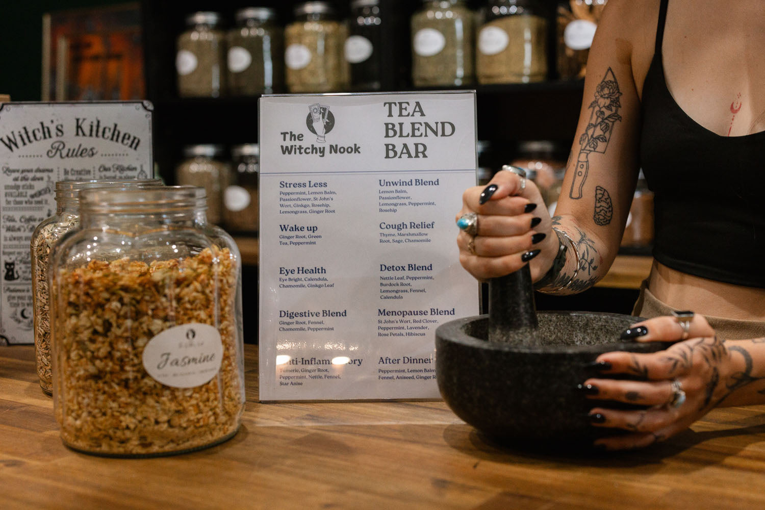 Person using a mortar and pestle at a tea blend bar at The Witchy Nook with jars and signs in the background.