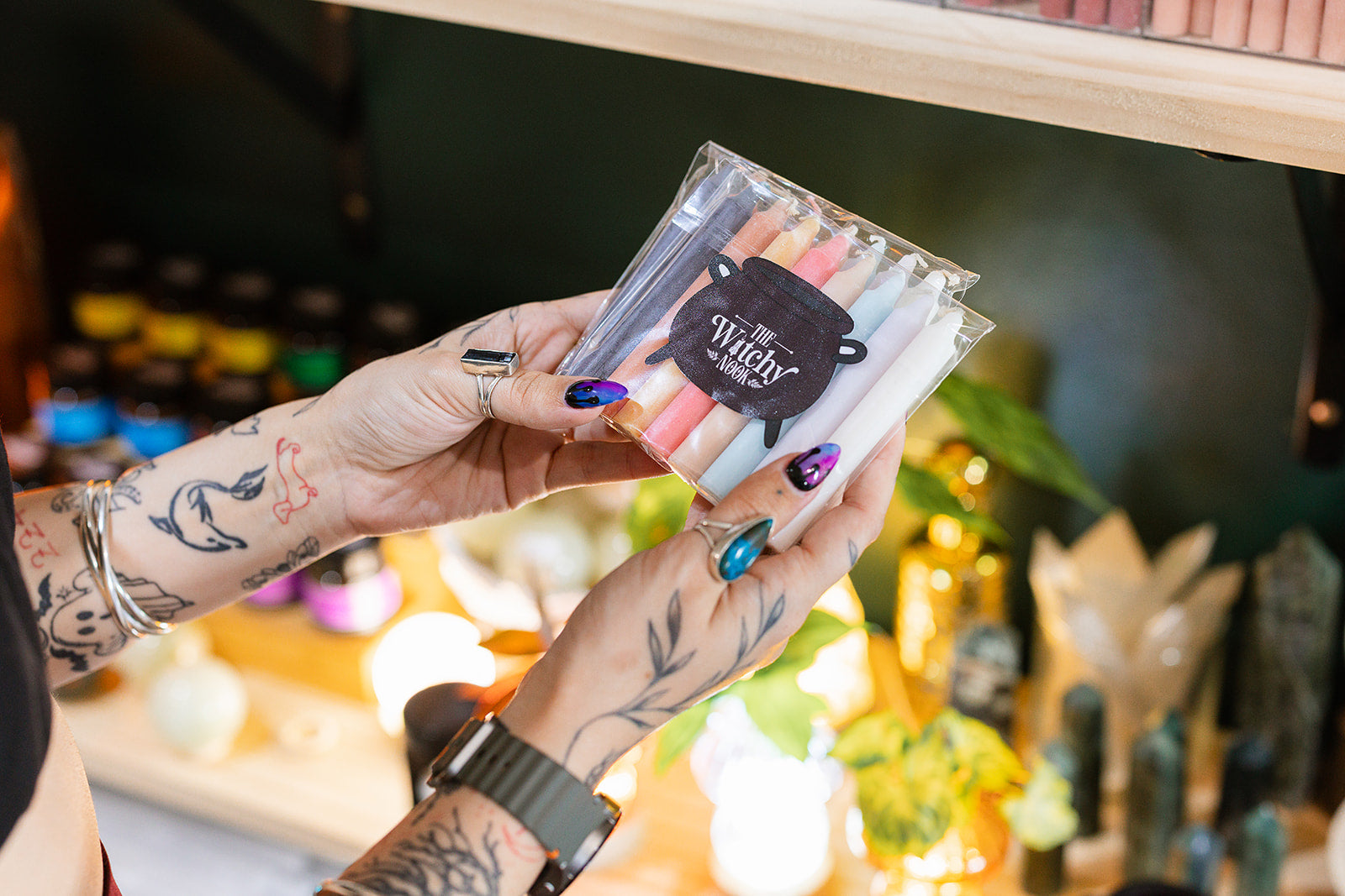 In this image you see a person holding a pack of spell candles from a store called "The Witchy Nook". The pack contains several small, colourful taper candles, often used in rituals or intention setting.