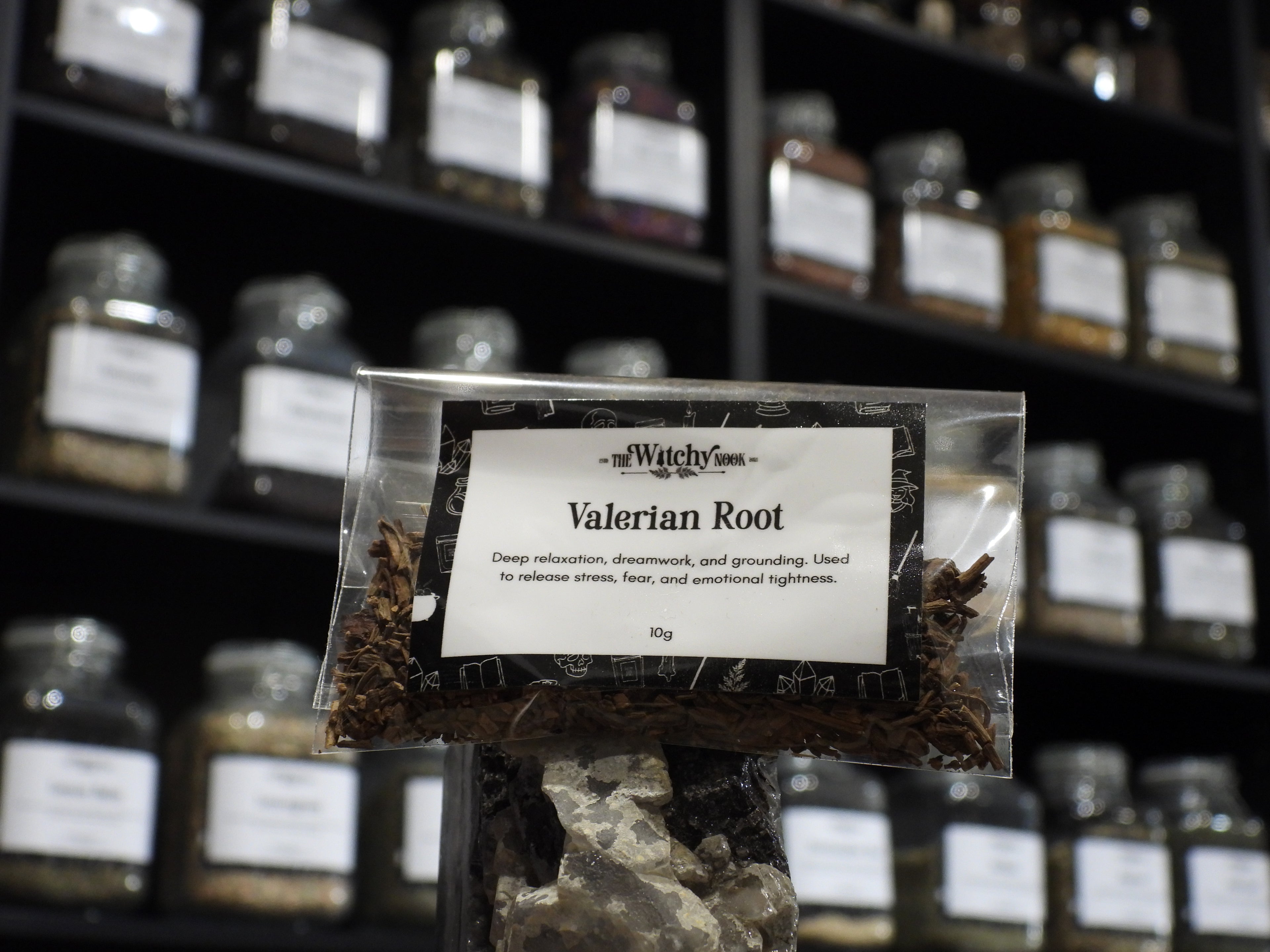 Packaged product labelled ‘Valerian Root' by 'Witchy Nook' sitting on a crystal with an apothecary of herb jars blurred in the background.