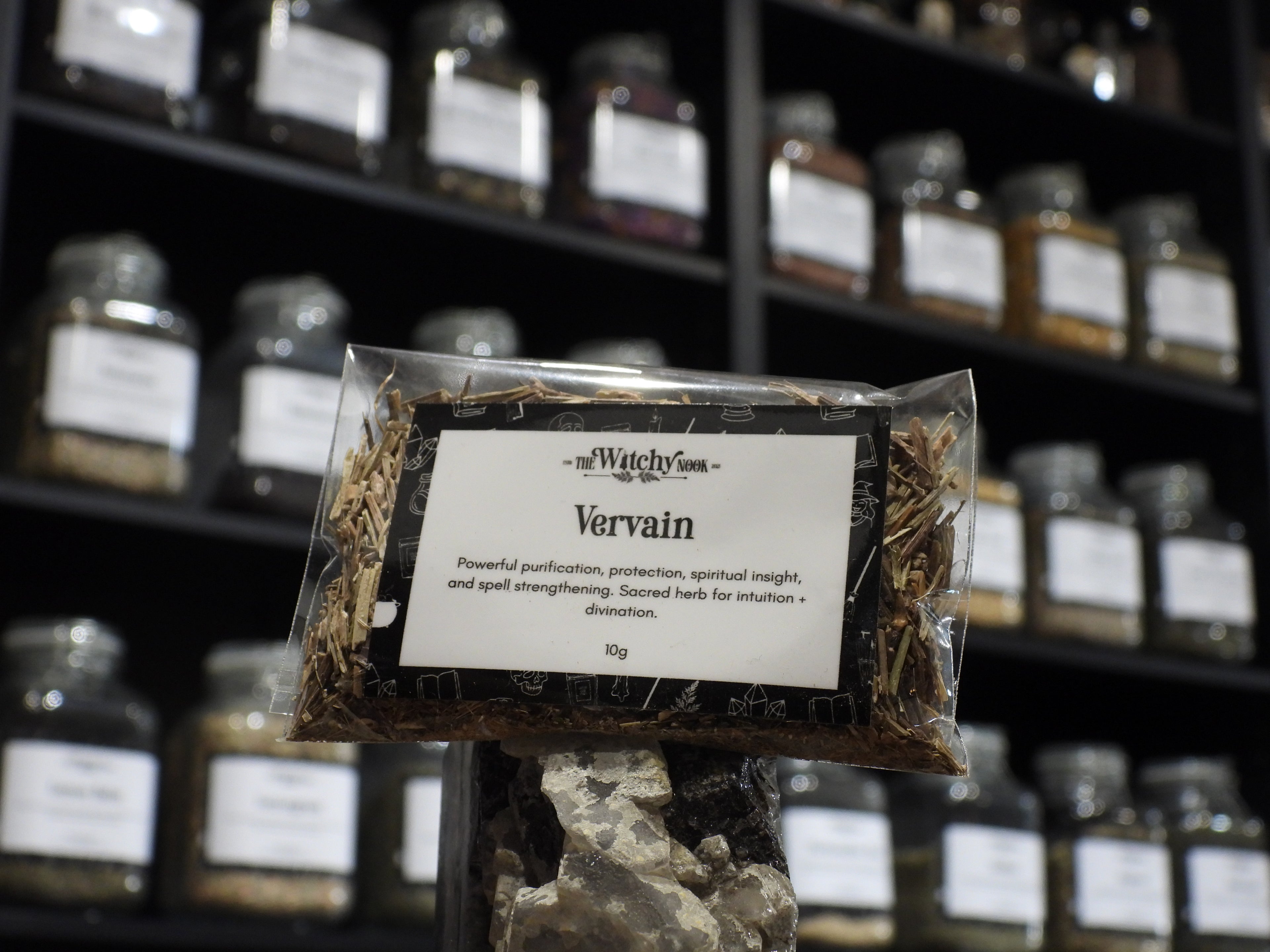 Packaged product labelled ‘Vervain' by 'Witchy Nook' sitting on a crystal with an apothecary of herb jars blurred in the background.