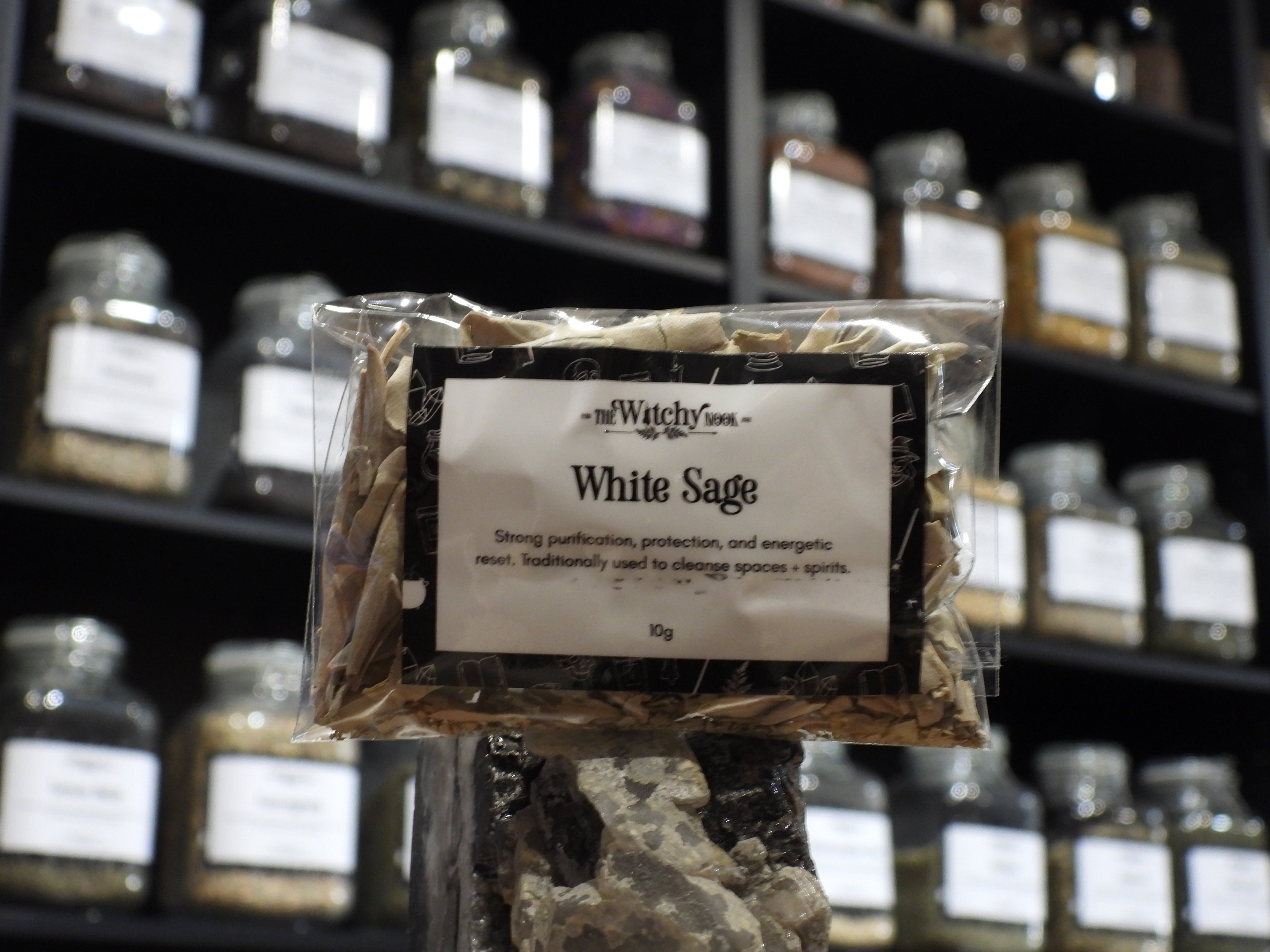 Packaged product labelled ‘White sage' by 'Witchy Nook' sitting on a crystal with an apothecary of herb jars blurred in the background.