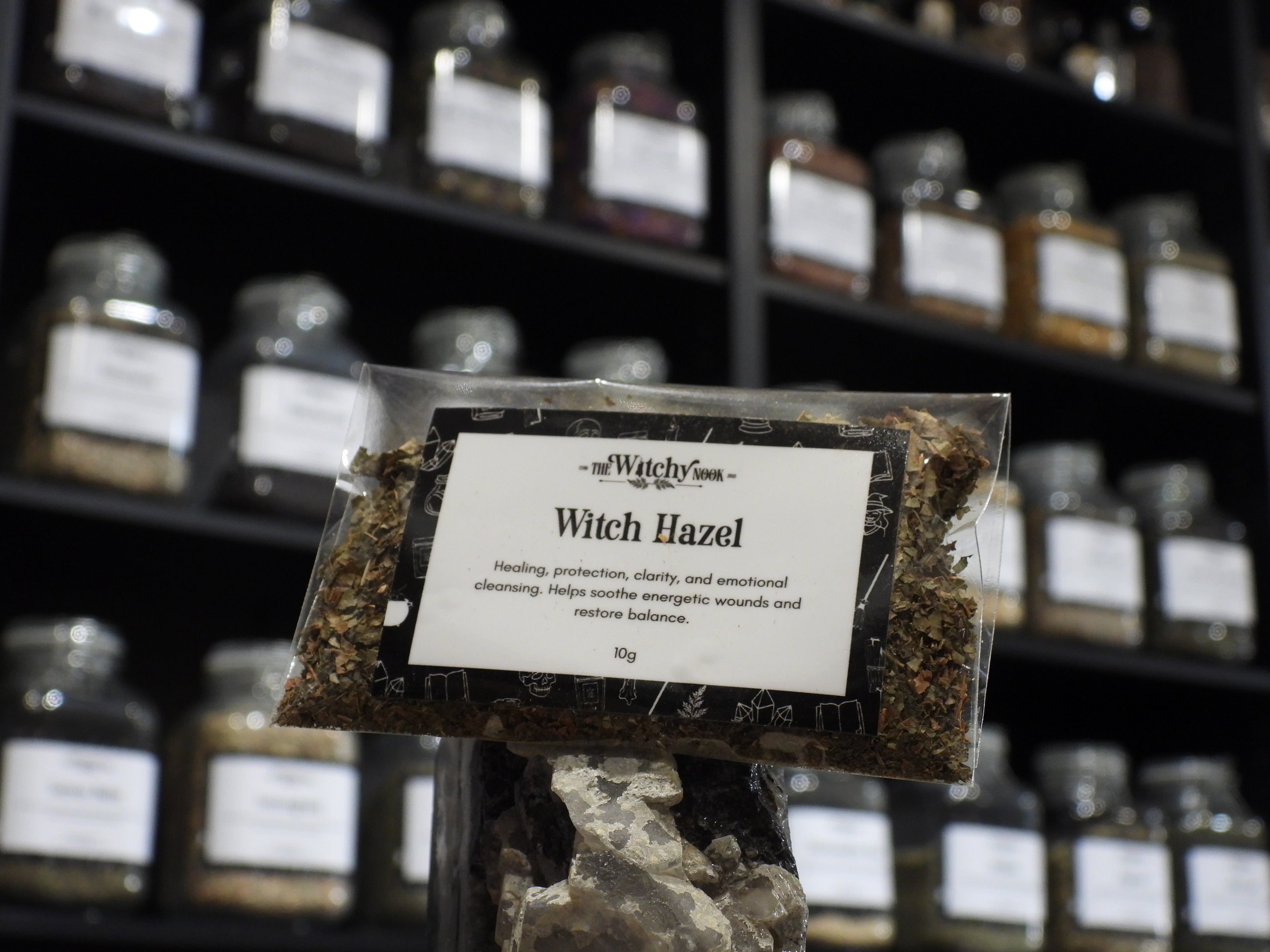 Packaged product labelled ‘Witch Hazel' by 'Witchy Nook' sitting on a crystal with an apothecary of herb jars blurred in the background.