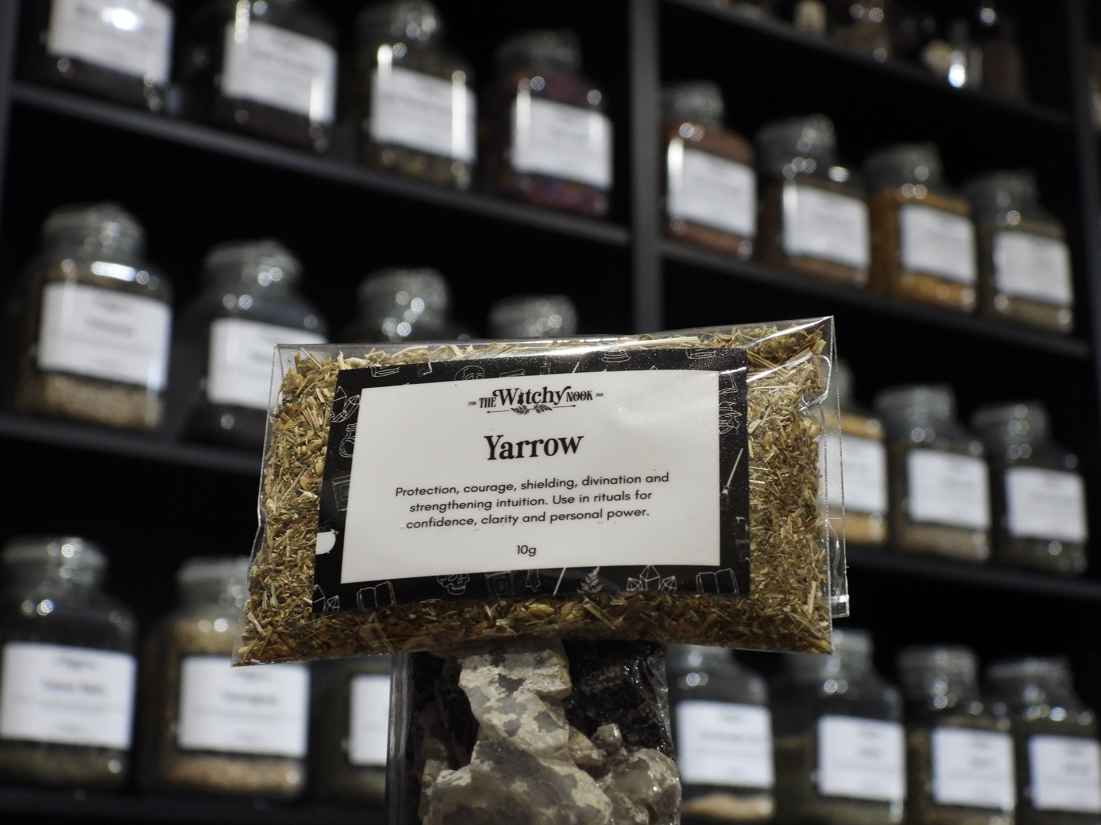 Packaged product labelled ‘Yarrow' by 'Witchy Nook' sitting on a crystal with an apothecary of herb jars blurred in the background.