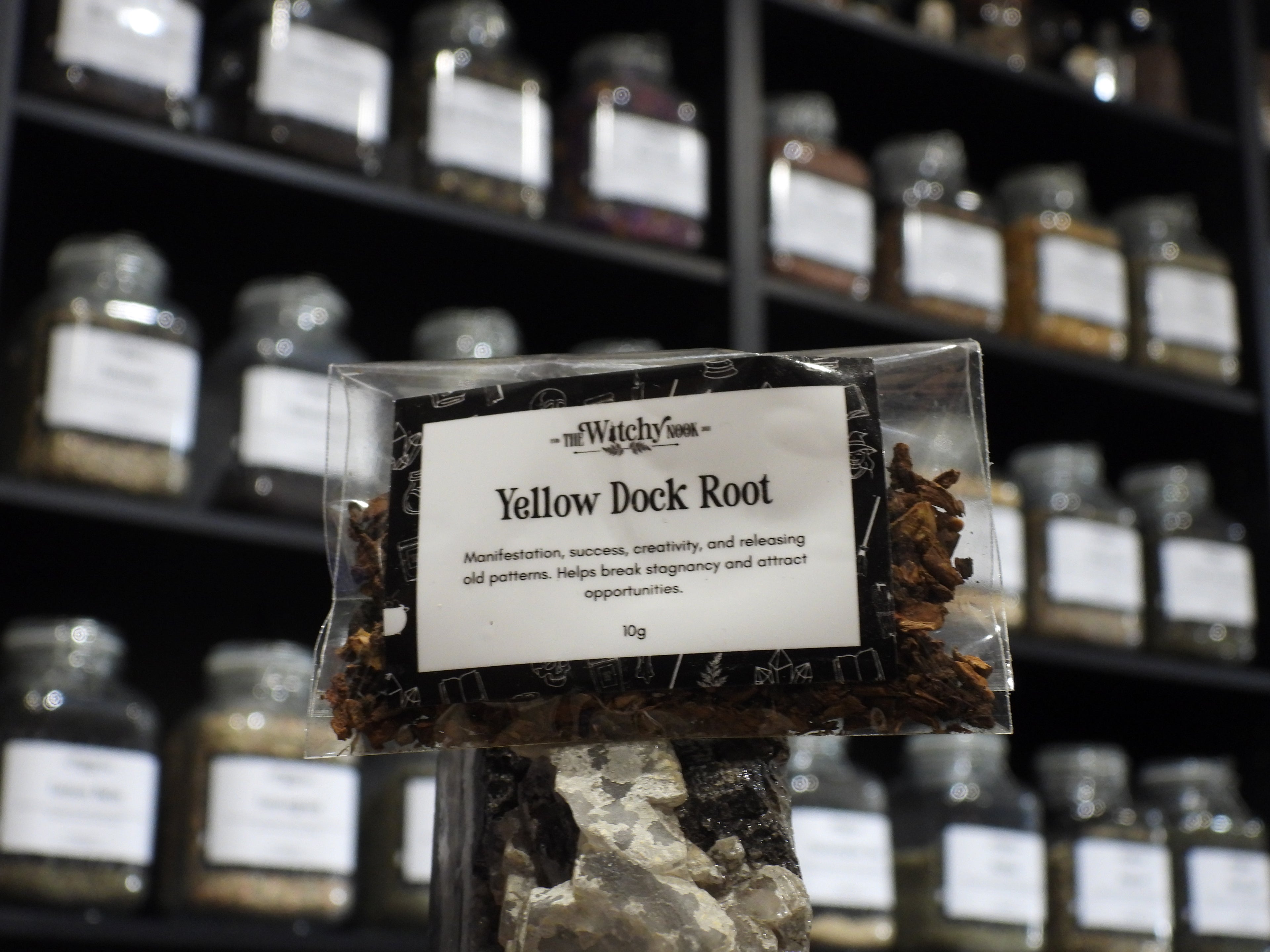 Packaged product labelled ‘Chilli Flakes' by 'Witchy Nook' sitting on a crystal with an apothecary of herb jars blurred in the background.